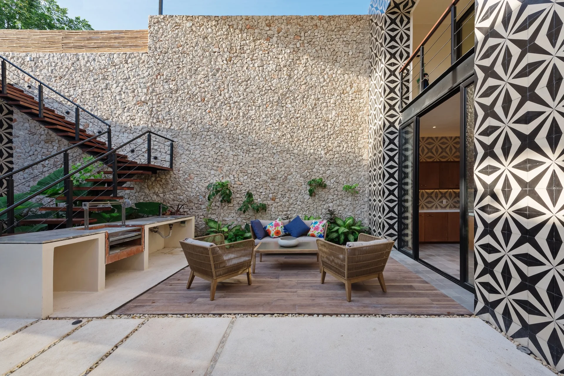 An outdoor patio features a large natural stone wall, wood-look decking with wicker furniture, a concrete outdoor kitchen, and geometric star-patterned tiles.