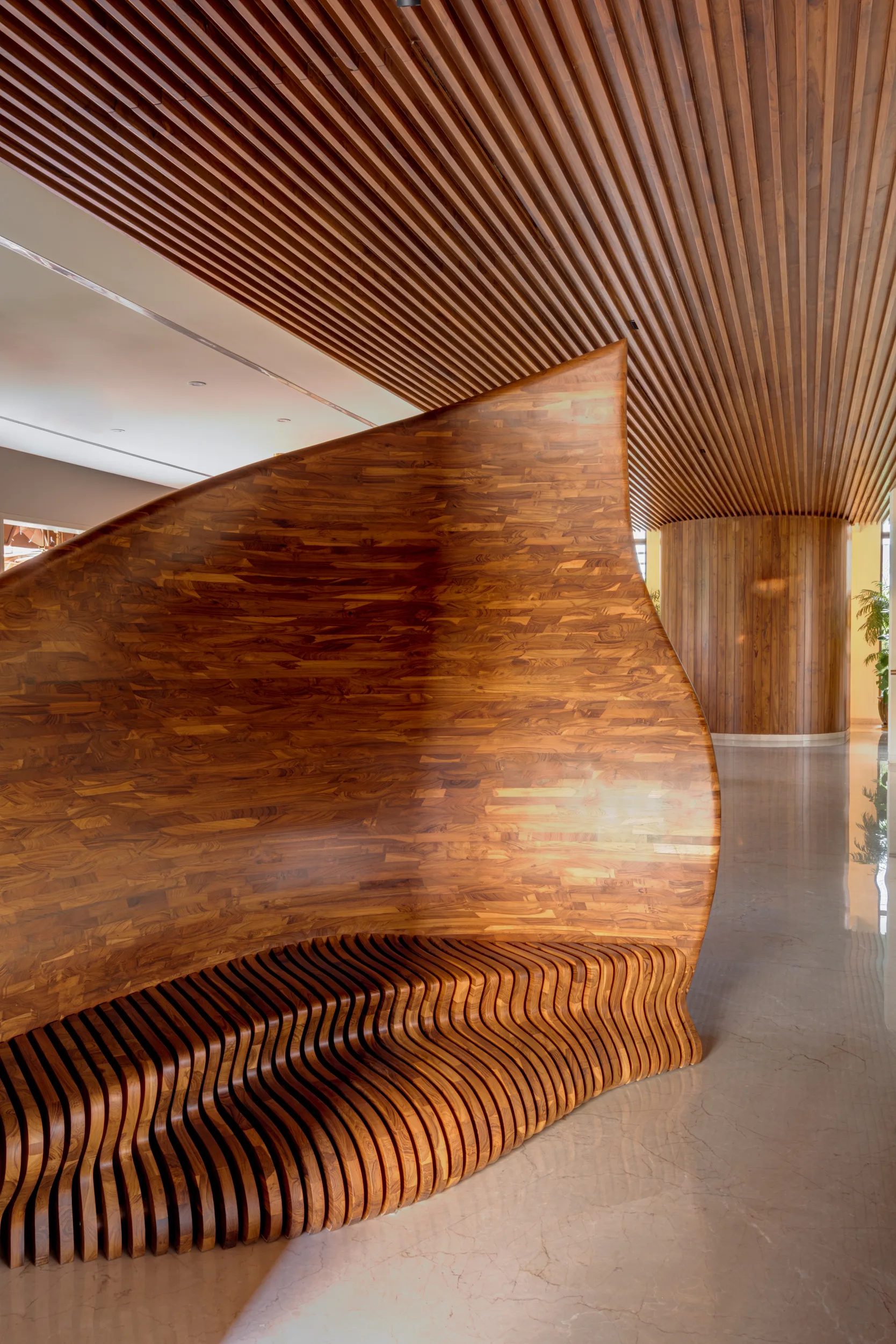 A large sculptural wooden parametric bench sits beneath a matching slatted wood ceiling on a polished beige stone floor.