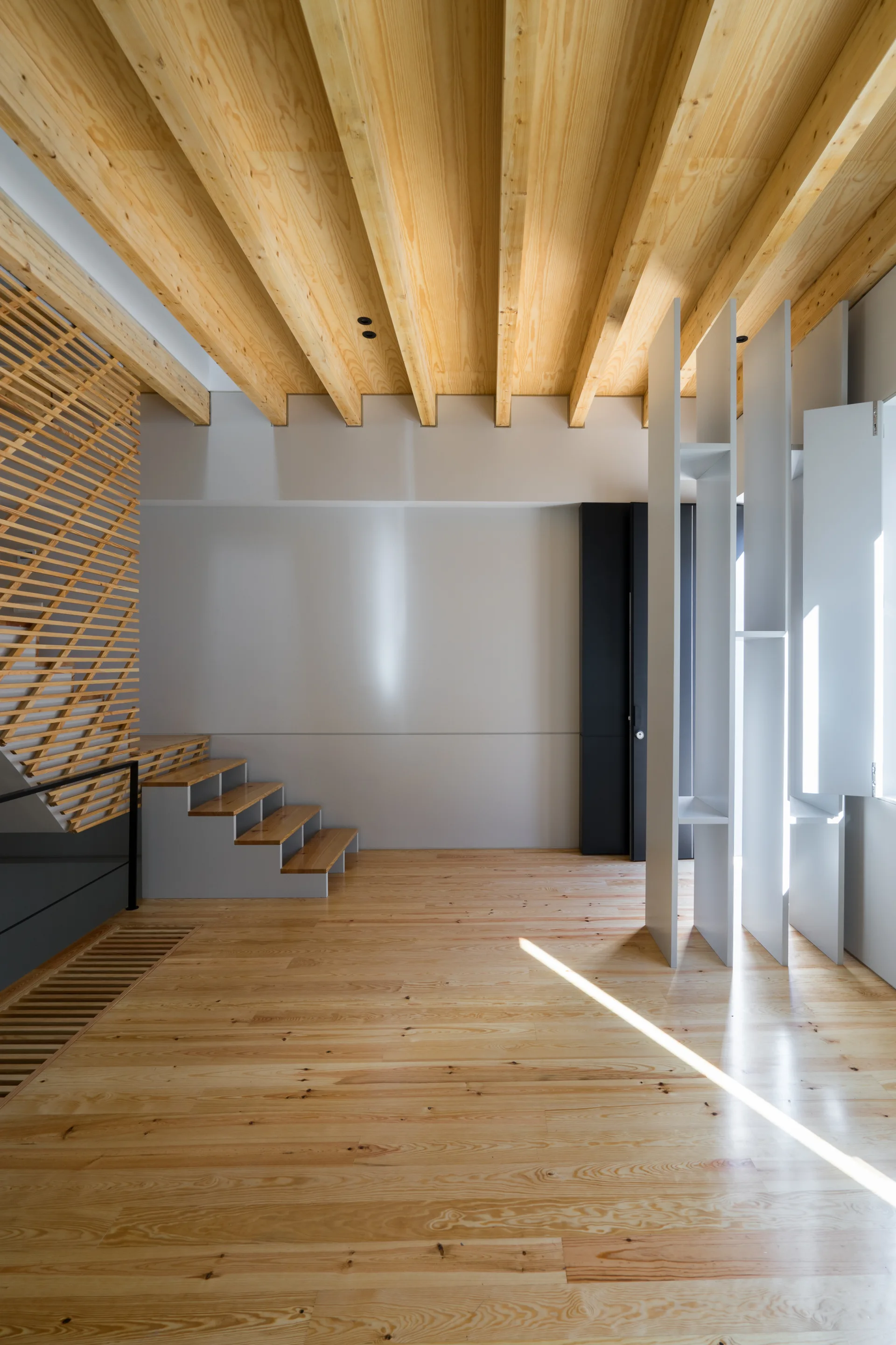 A minimalist entryway features light wood floors, matching exposed ceiling beams, a wooden staircase with a slatted screen, and a white vertical room divider.
