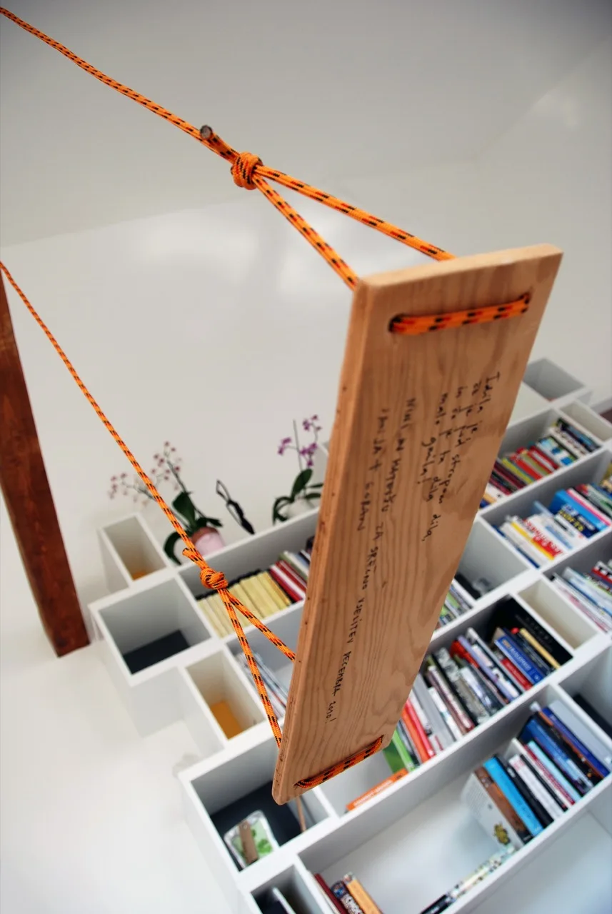 A wooden swing seat with bright orange ropes hangs in front of a white geometric bookshelf filled with colorful books.