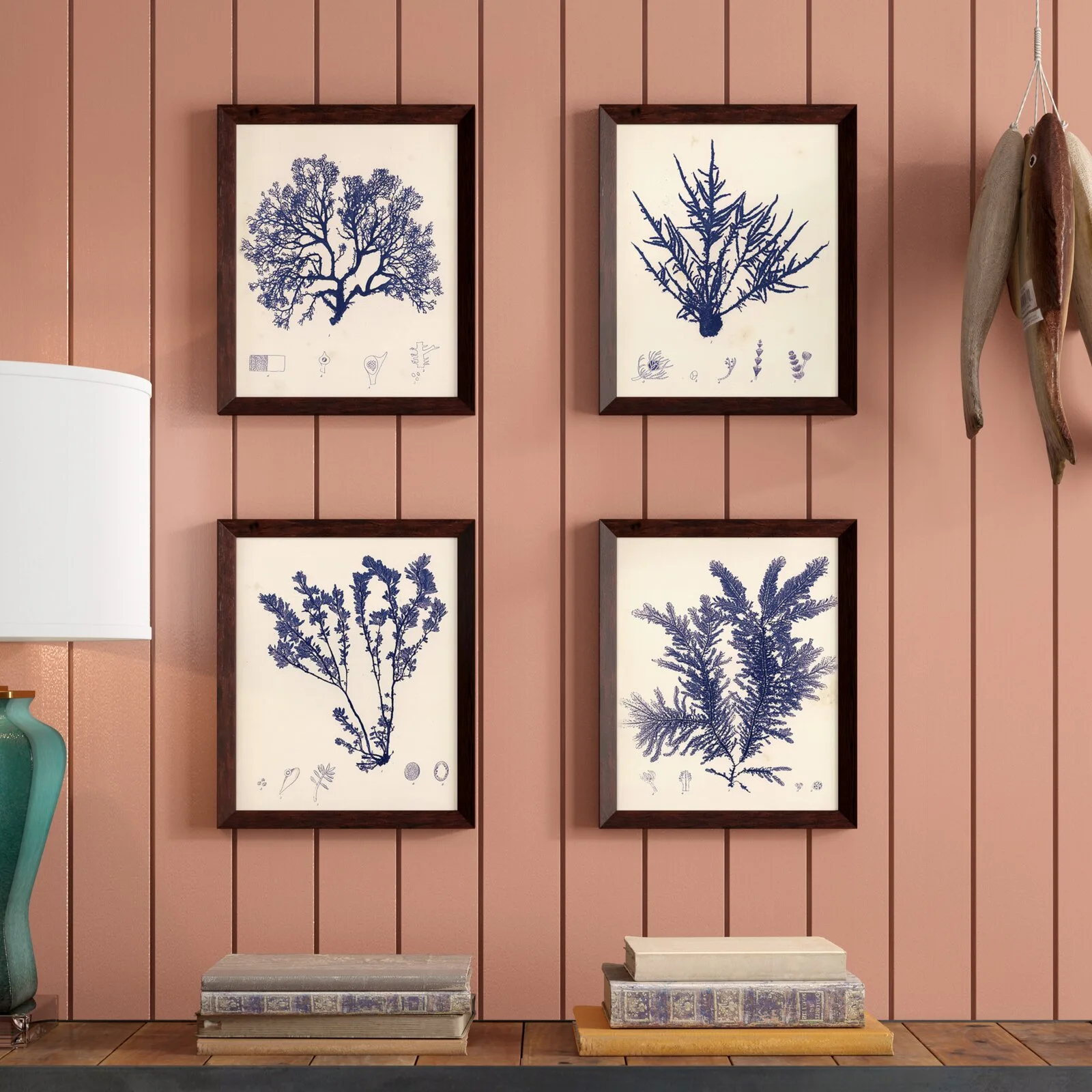 Four symmetrically framed blue seaweed prints hang on a salmon pink vertical paneled wall above stacked books and a lamp.
