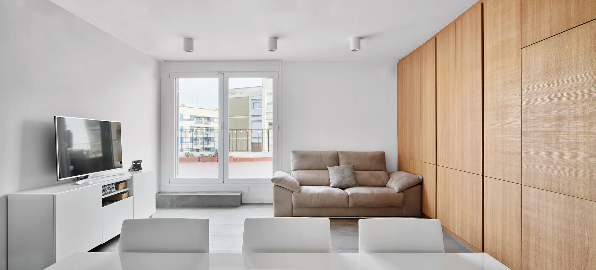 A minimalist living room features a floor-to-ceiling wood storage wall, a beige sofa, and a white media console with a TV.