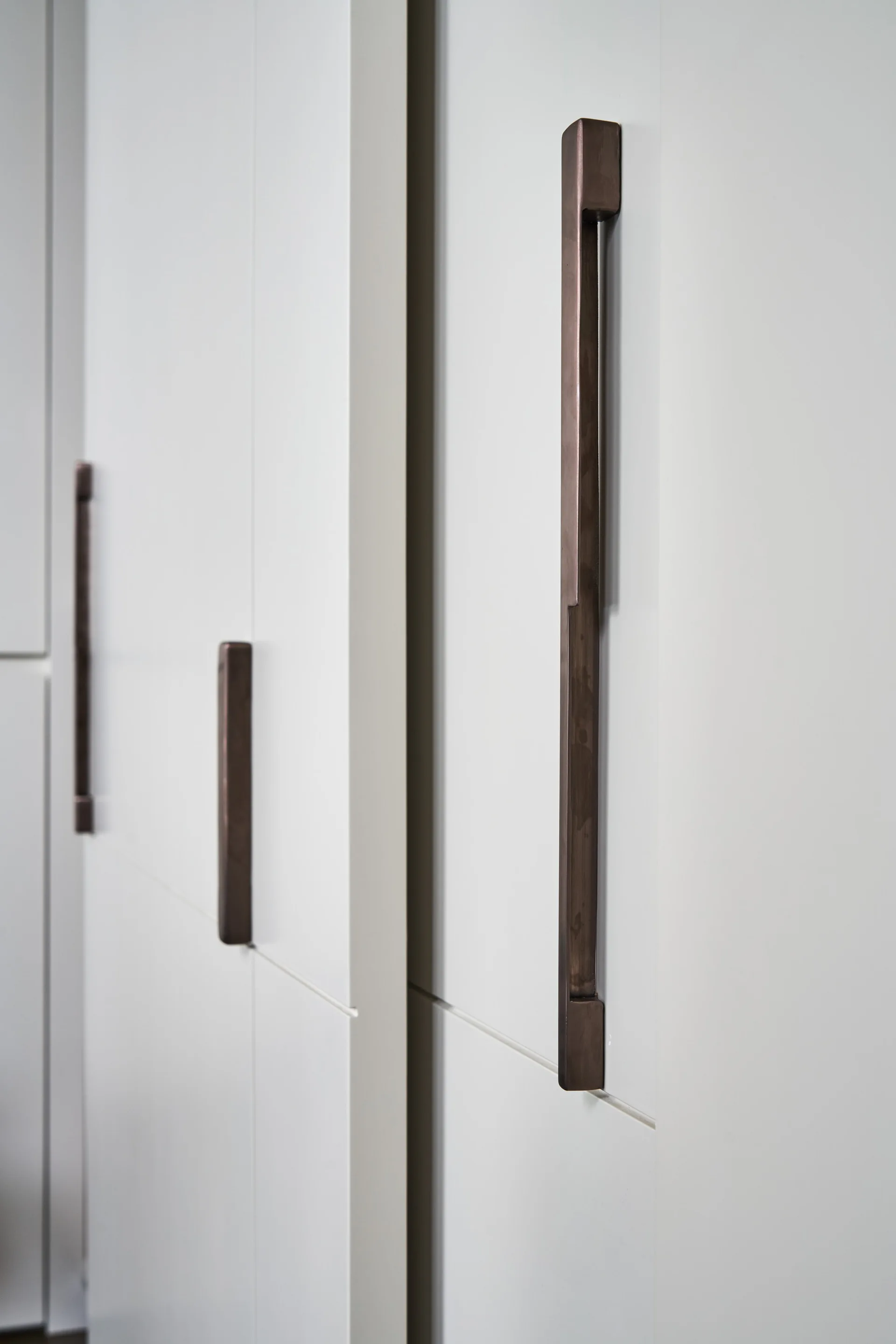 A close-up view of flat white cabinet doors fitted with long, vertical dark bronze metal bar handles.