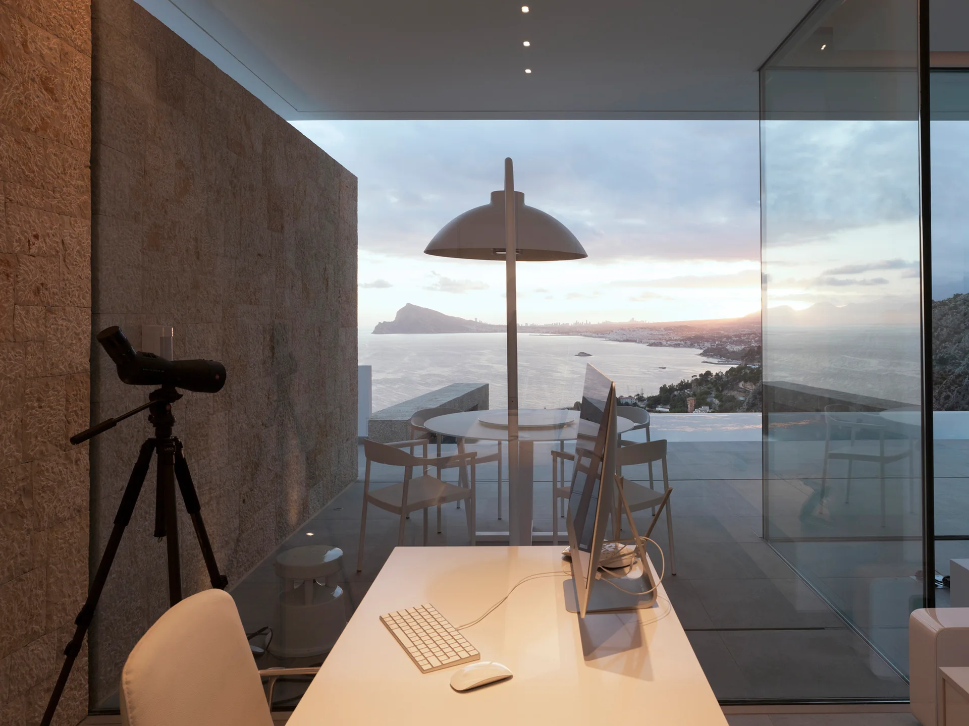 A modern home office with a desk and computer features a textured stone wall, a telescope, and a glass partition looking out to a sunset sea view.