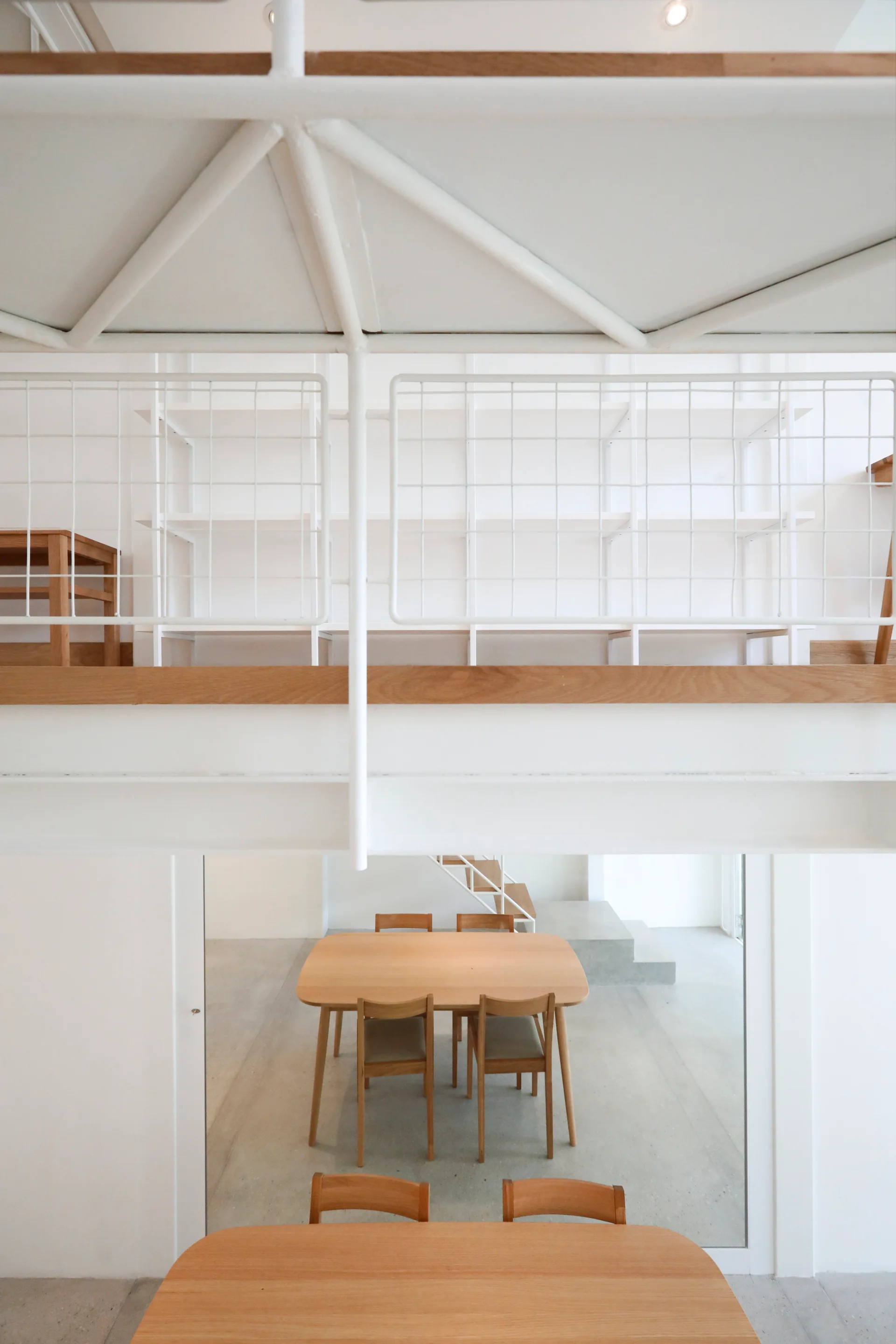 A minimalist dining area with light wood tables and chairs viewed from underneath a white mezzanine with a metal grid railing.