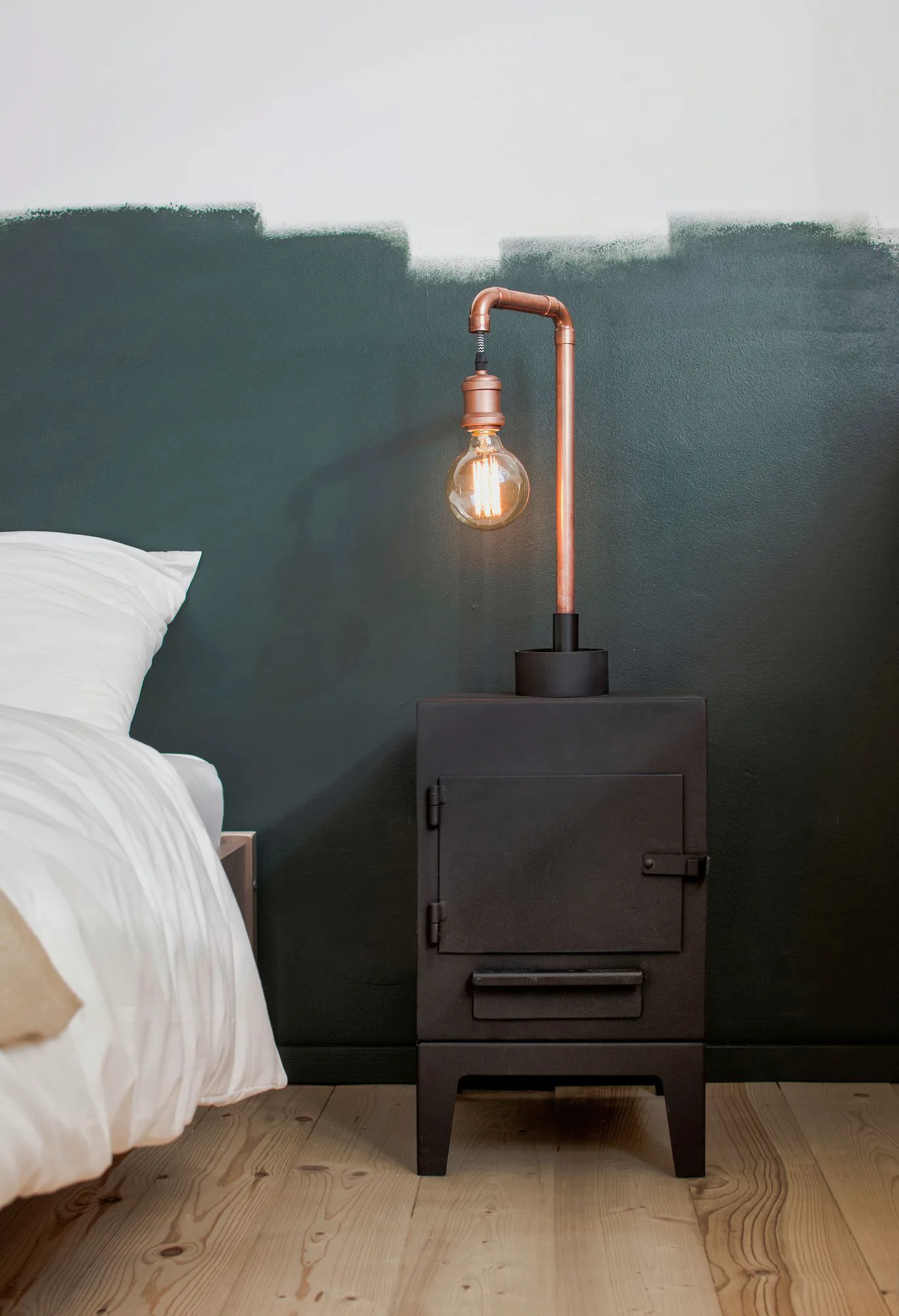 A copper pipe desk lamp sits on a black metal nightstand in a bedroom with a dark green accent wall and light wood flooring.