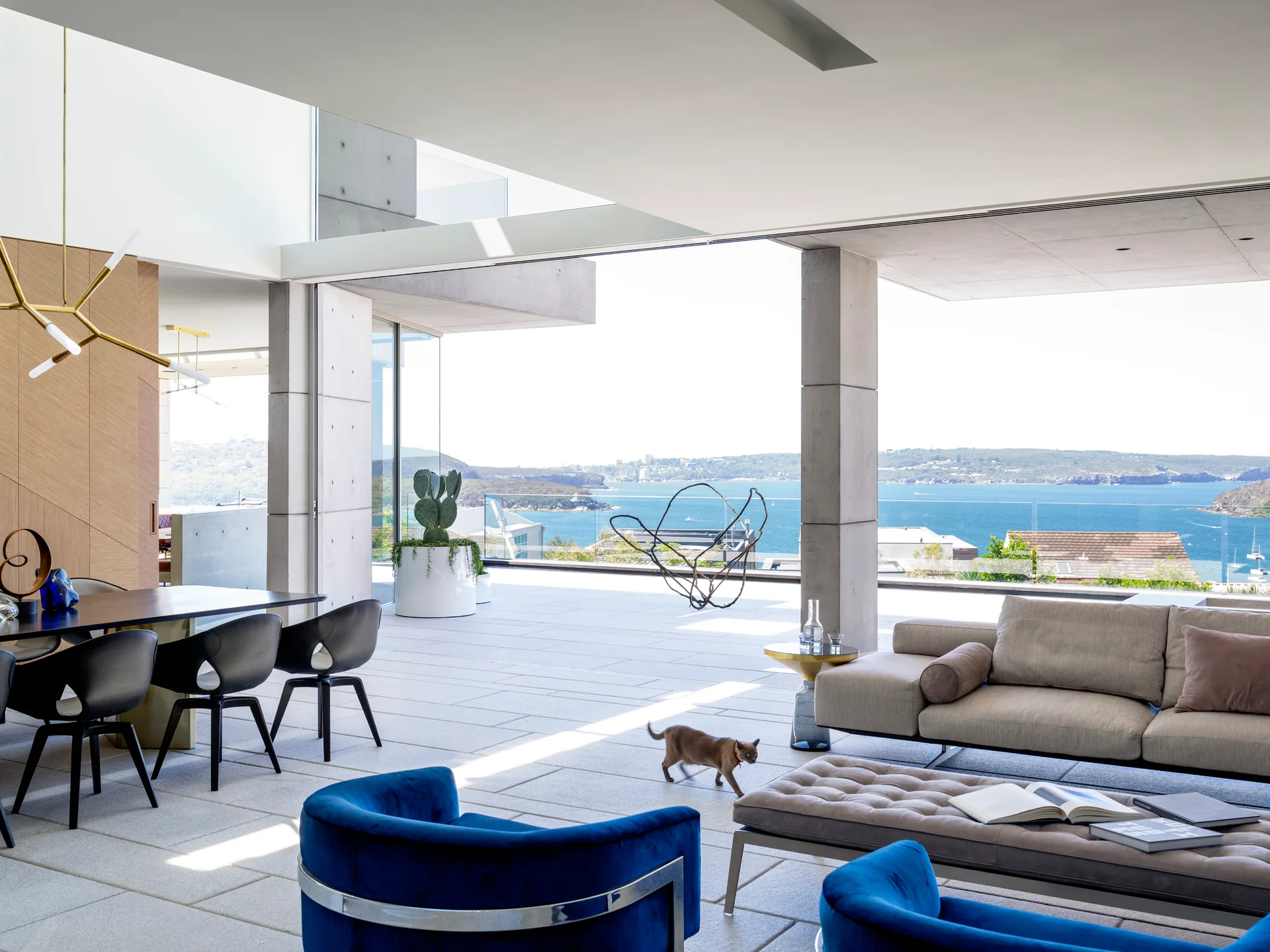 A modern open-plan living room with beige and blue furniture opens onto a stone terrace with concrete pillars and a scenic ocean view.
