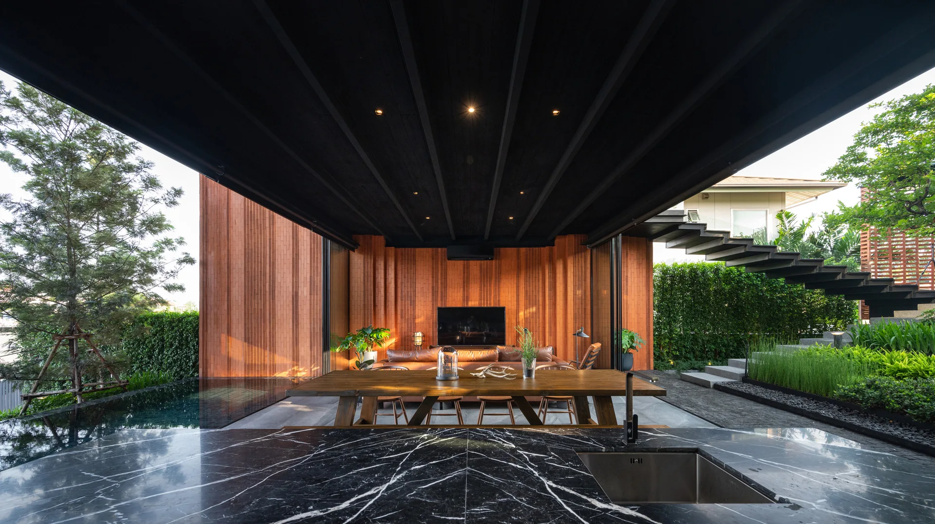 A covered open-air patio features a wood dining table, a brown leather sofa against wood slat walls, a dark ceiling, and an adjacent swimming pool.
