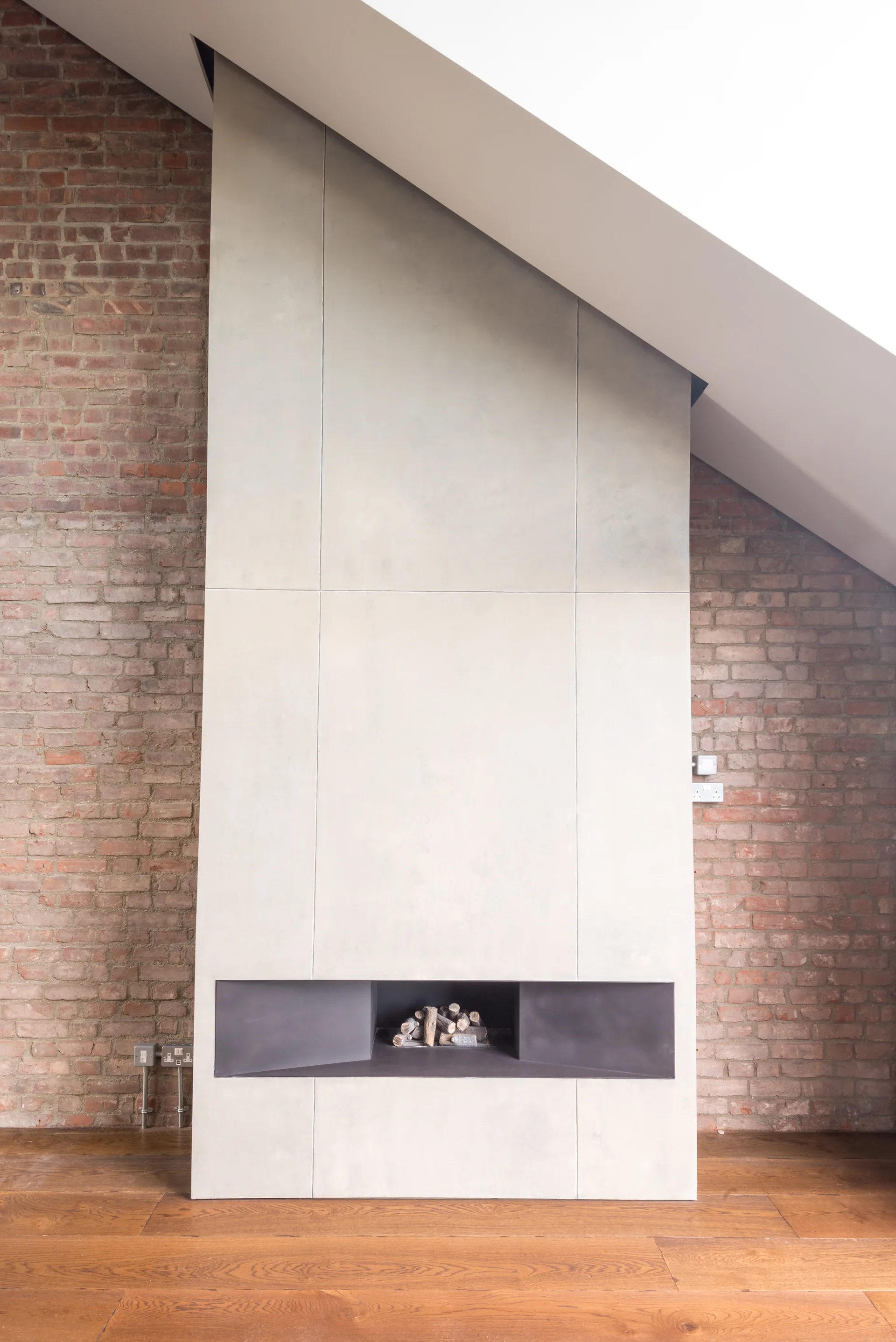A modern off-white paneled fireplace chimney breast stands against an exposed red brick wall under a sloped ceiling with hardwood floors below.