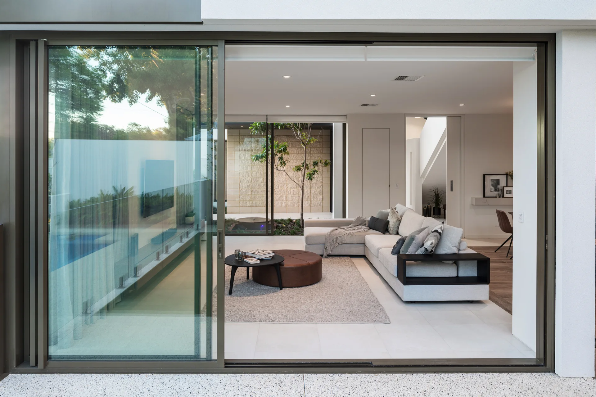 A modern living room features a light grey sectional sofa and a brown leather ottoman next to large sliding glass doors that open to an outdoor courtyard.