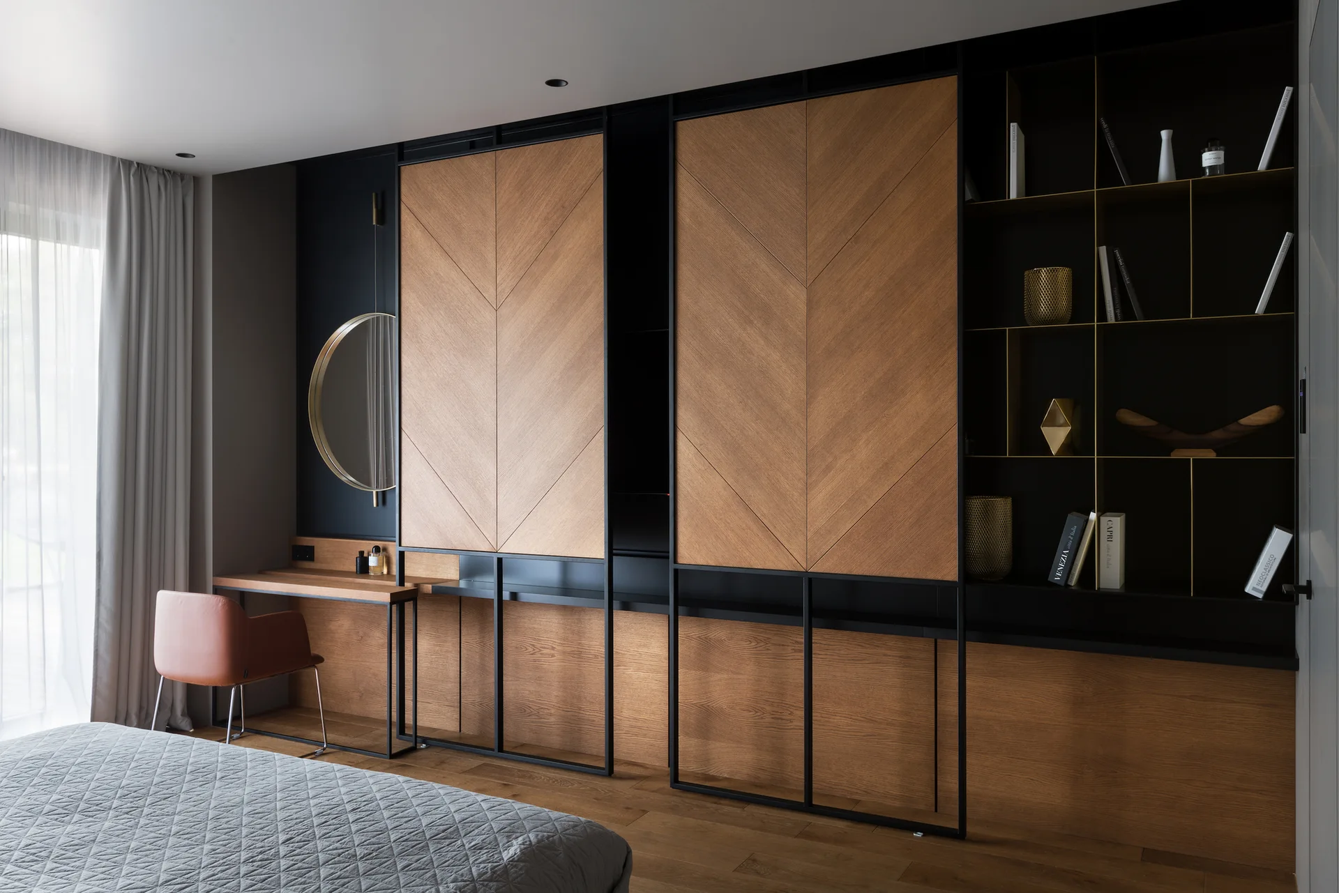 A modern bedroom features a custom wood and black metal cabinetry wall with chevron panels, an integrated vanity desk, and a grey quilted bed.