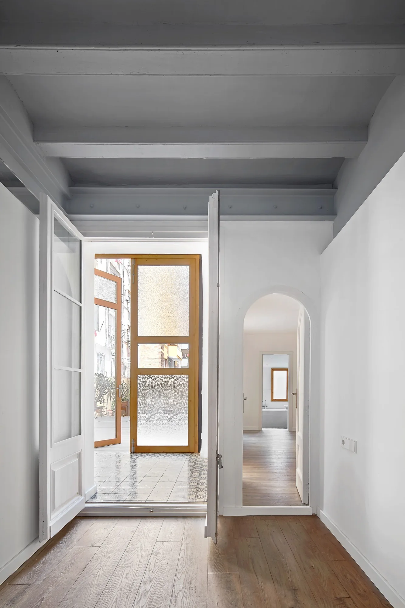 A bright interior hallway with wood flooring features grey ceiling beams, an arched doorway, and double doors opening onto a patterned tile patio.