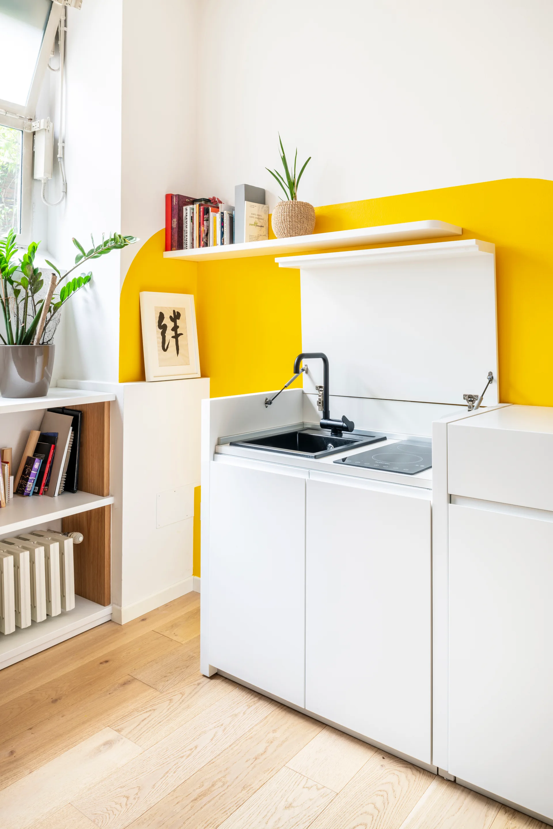 A compact white kitchenette with a black sink stands against a white wall decorated with a large curved yellow painted shape.