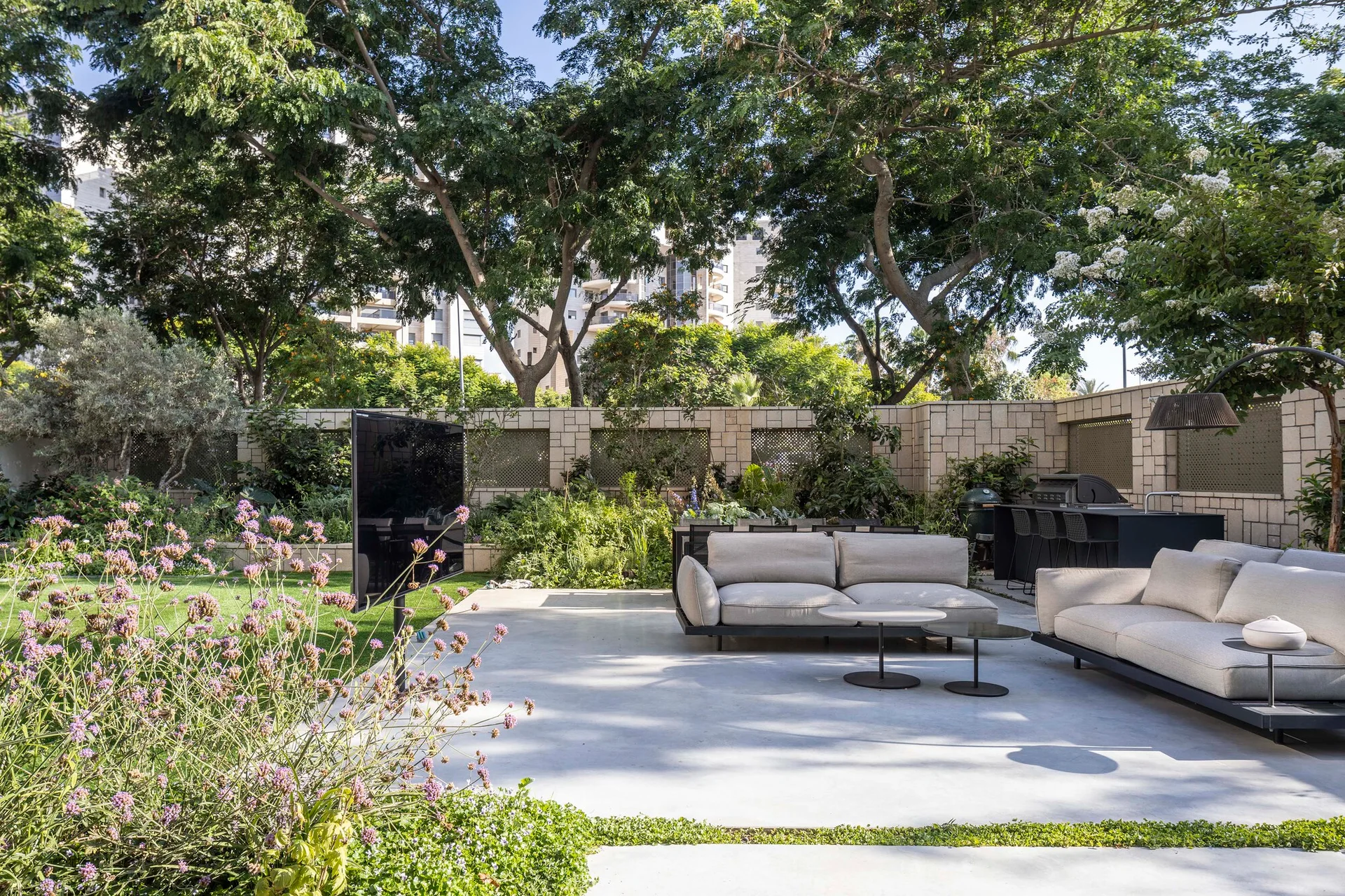 Two light gray outdoor modular sofas sit on a smooth concrete patio next to a manicured lawn, framed by a stone block wall and tall trees.