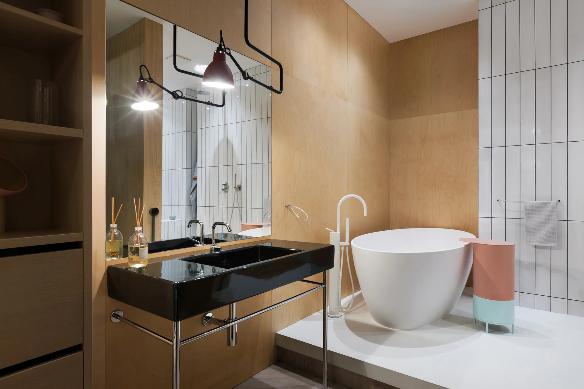 A contemporary bathroom features a black vanity sink, light wood wall panels, vertical white tiles, and a white freestanding tub.