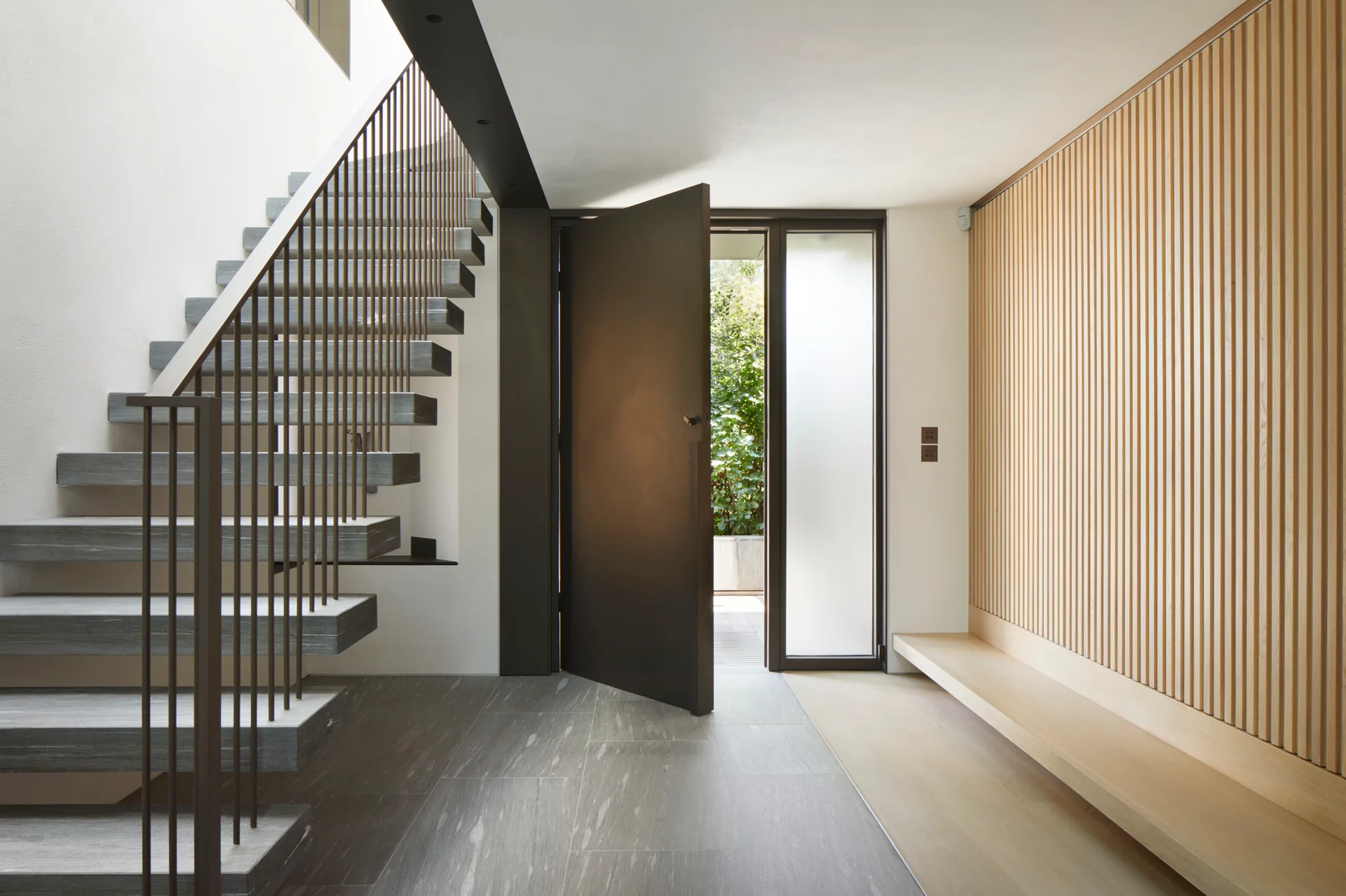 A modern foyer features an open-riser stone staircase, a dark open pivot door, and a light wood slat wall with an integrated bench.
