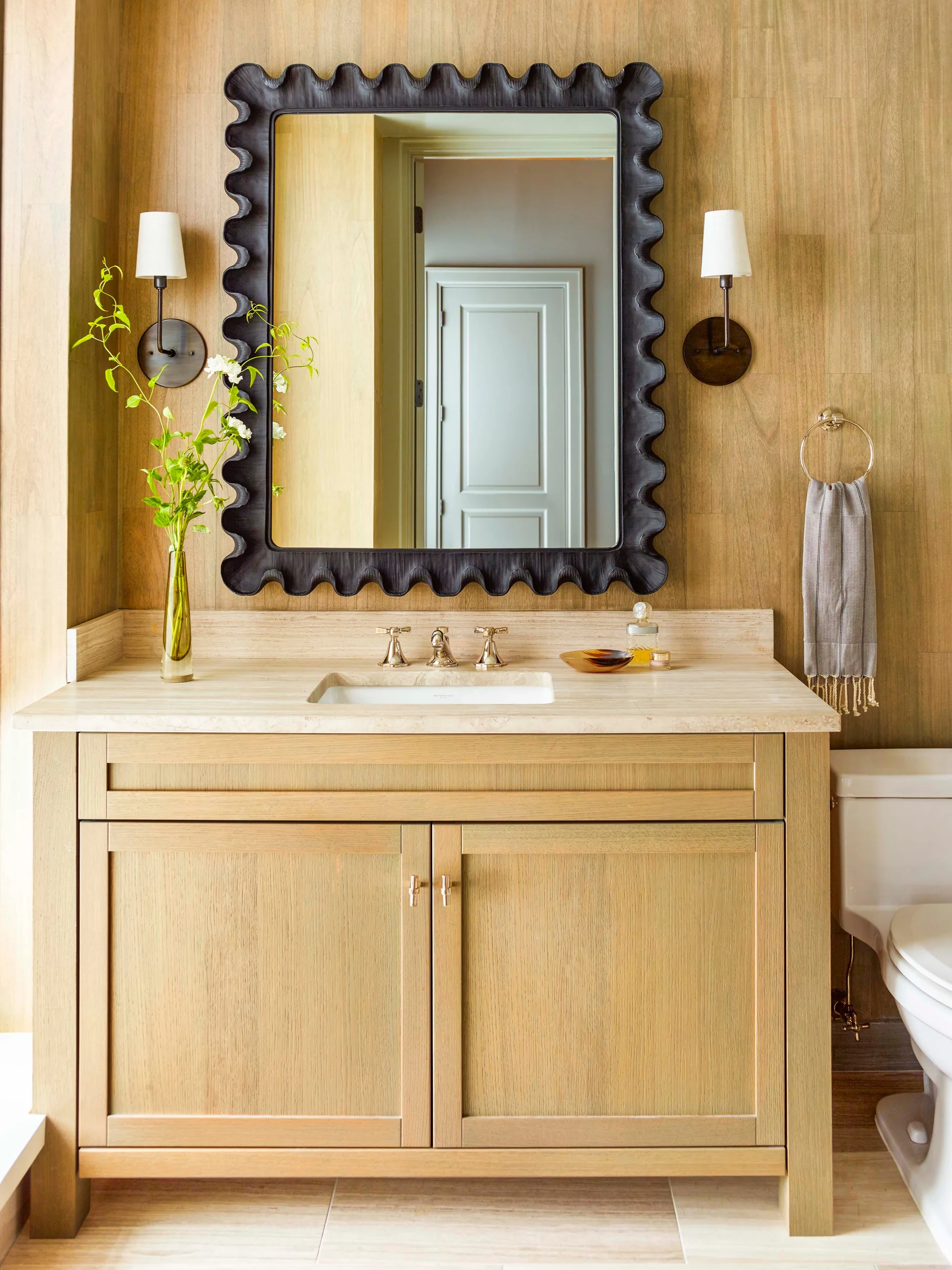 A light wood bathroom vanity with a stone countertop sits against a vertical wood plank wall featuring a black scalloped mirror and sconces.
