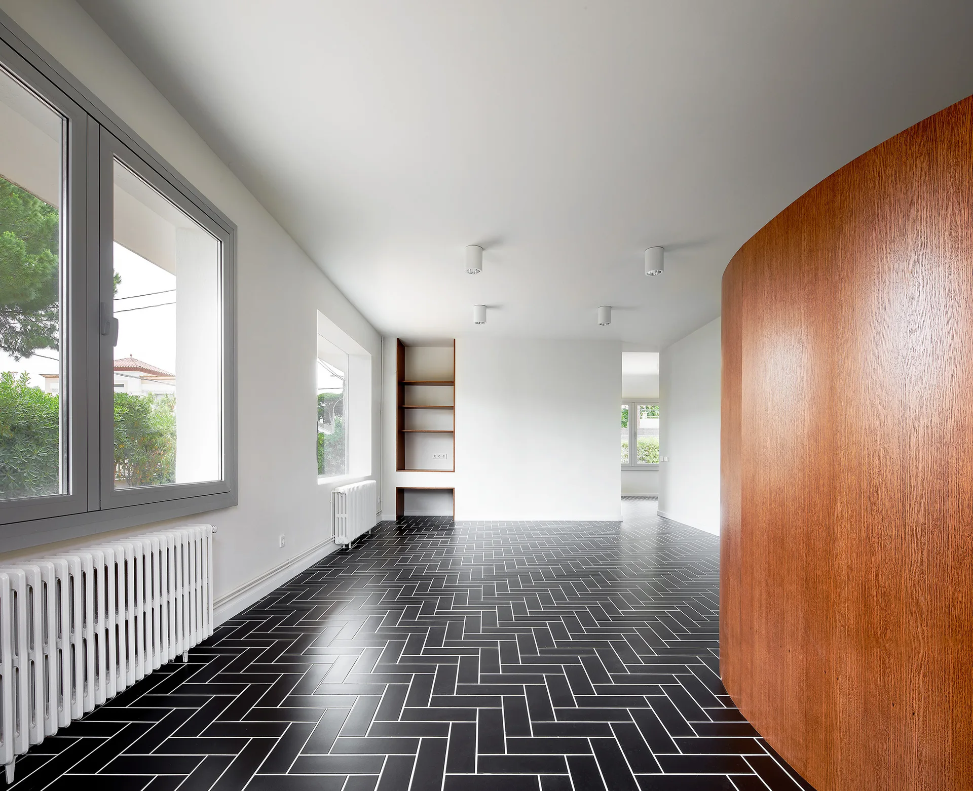 An empty room featuring black herringbone tile flooring, white walls with classic radiators, and a large curved wood partition wall.