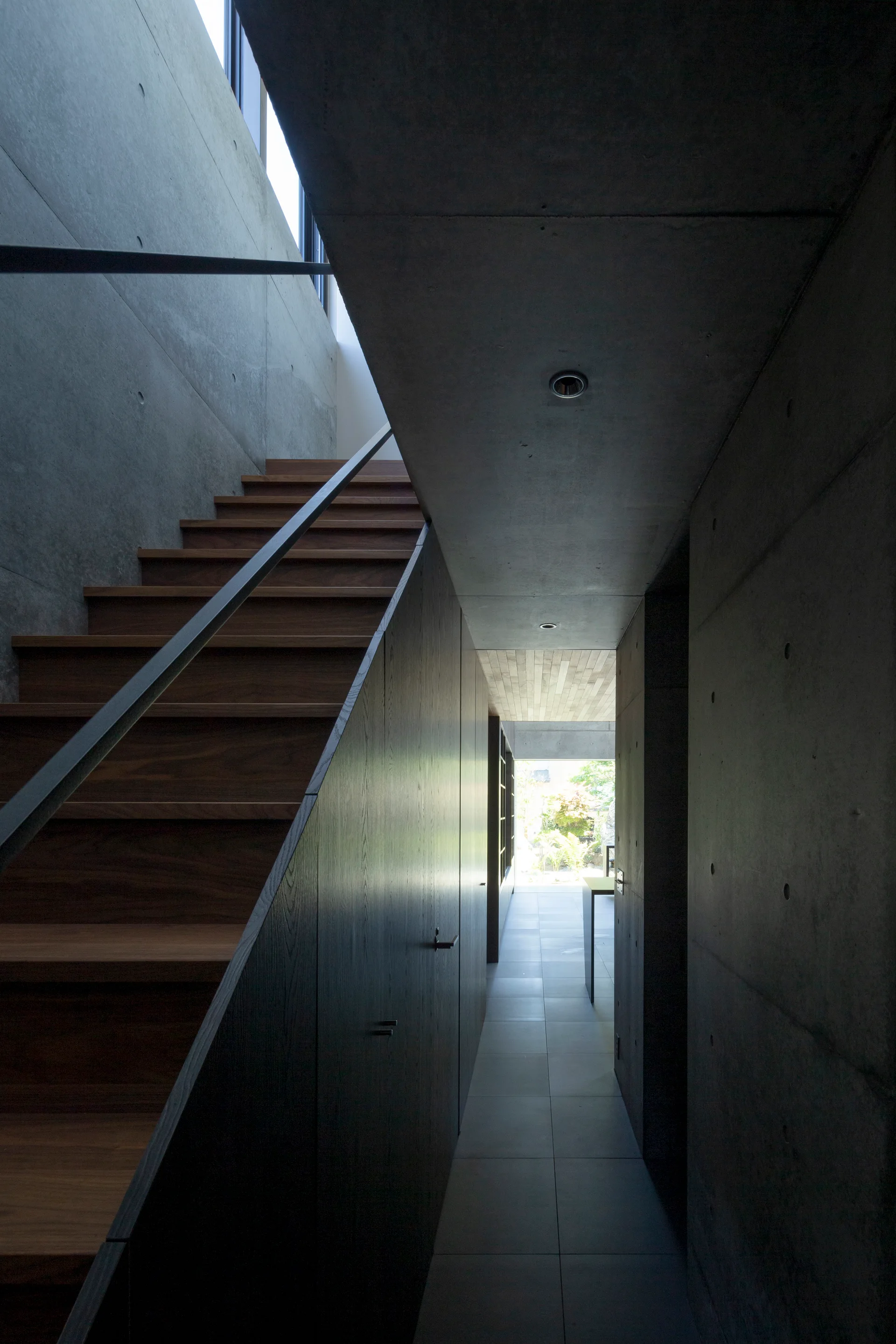 A minimalist hallway features raw concrete walls, warm wood stair treads, and dark wood paneled storage cabinets built underneath the stairs.