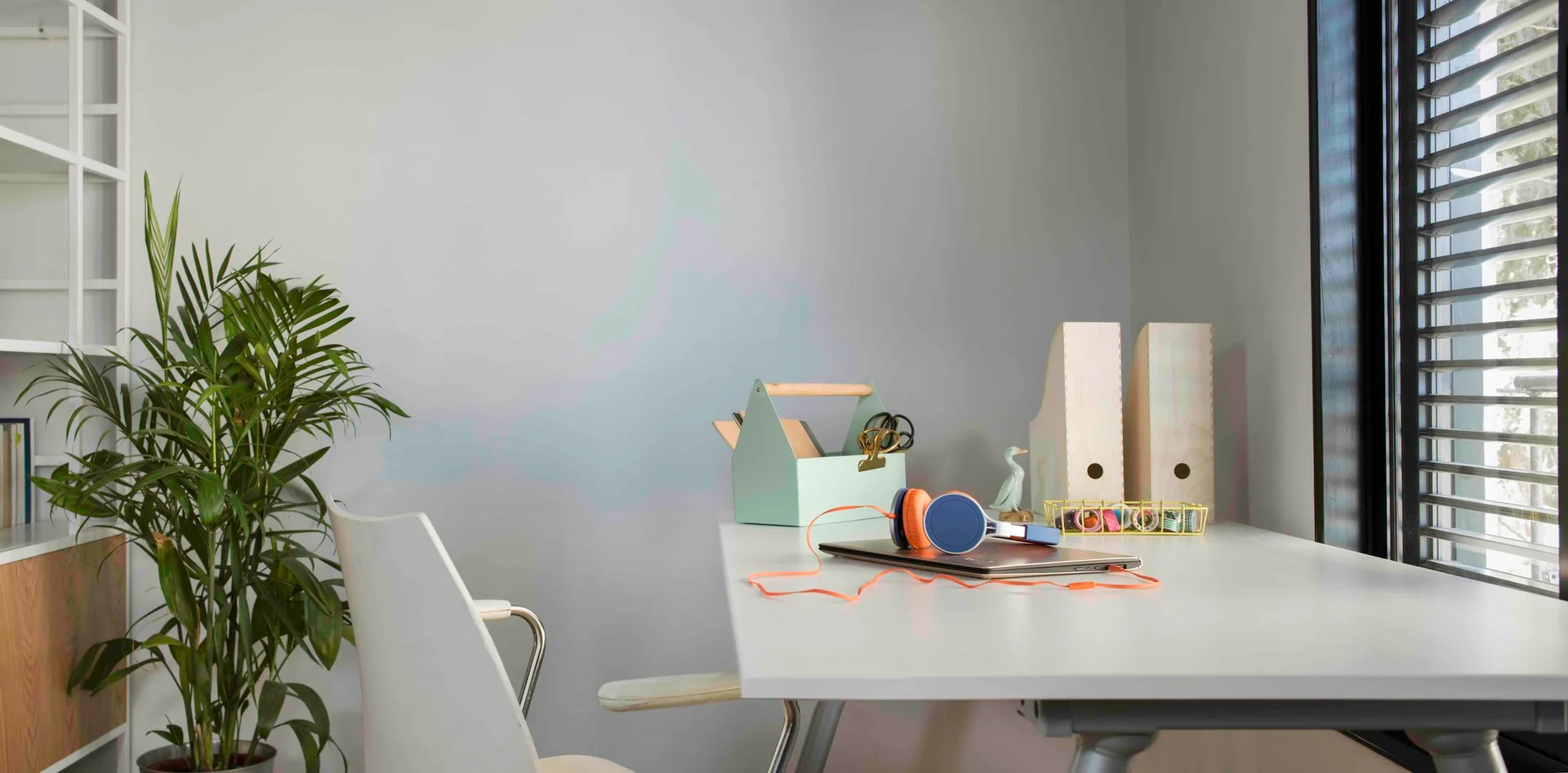 A minimalist white desk features a laptop, headphones, a green storage caddy, wooden file holders, and a potted plant near a window with black blinds.