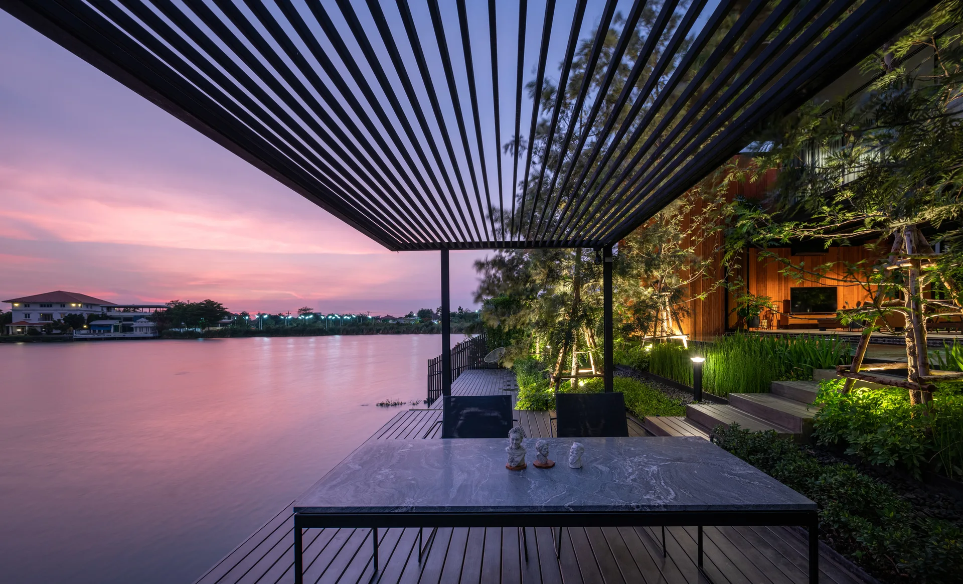 A wooden waterfront deck features a rectangular stone-topped dining table under a black slatted pergola at sunset.