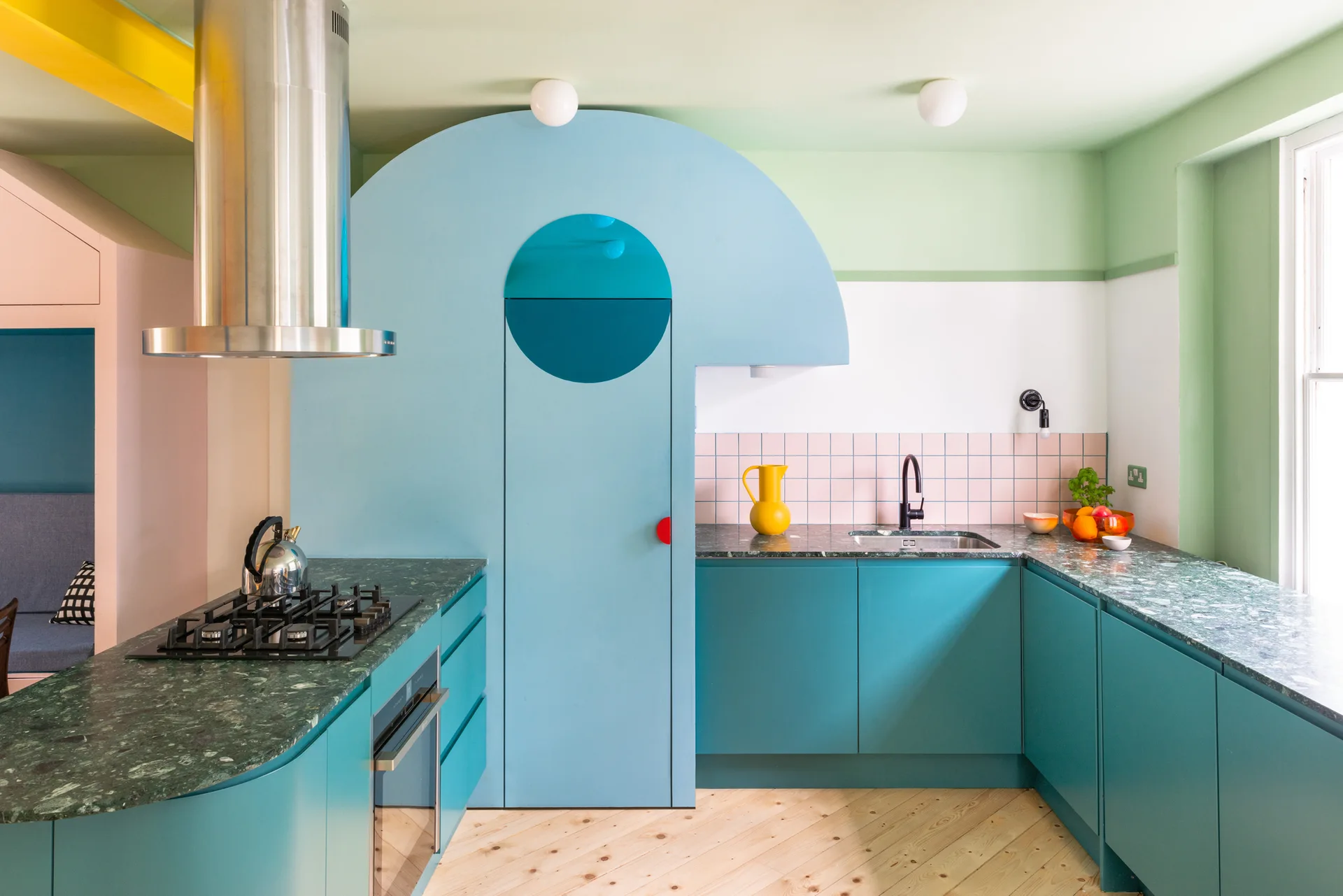 A colorful modern kitchen features teal cabinets, a green stone countertop, pink square tile backsplash, and a prominent curved blue panel over a pantry door.
