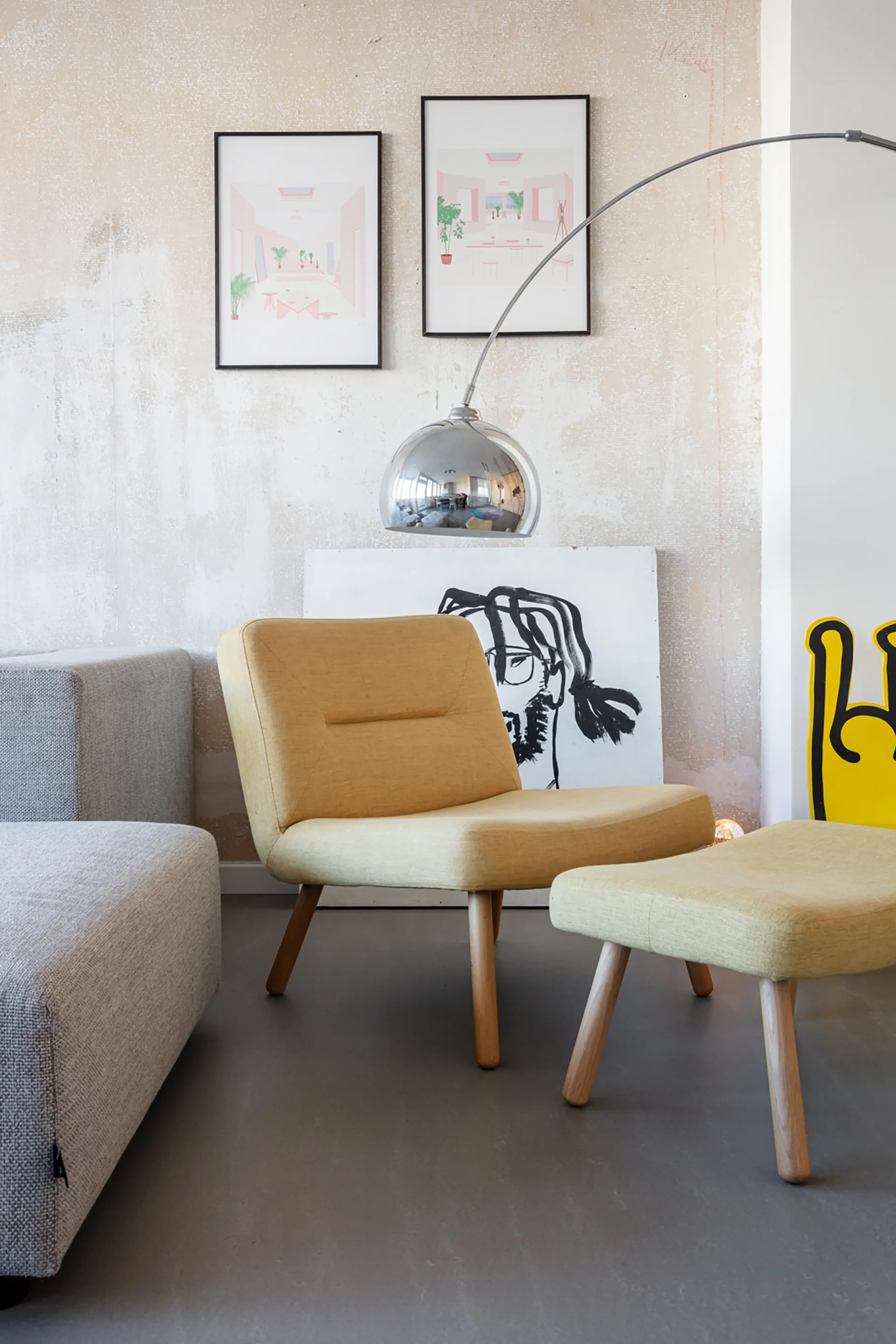 A living room corner features a yellow upholstered chair and matching ottoman, a grey sofa, and an arched metal floor lamp against a textured plaster wall.
