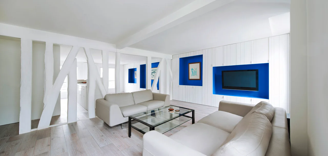 A modern white living room with off-white sofas, a glass coffee table, a structural timber partition, and deep blue wall niches.