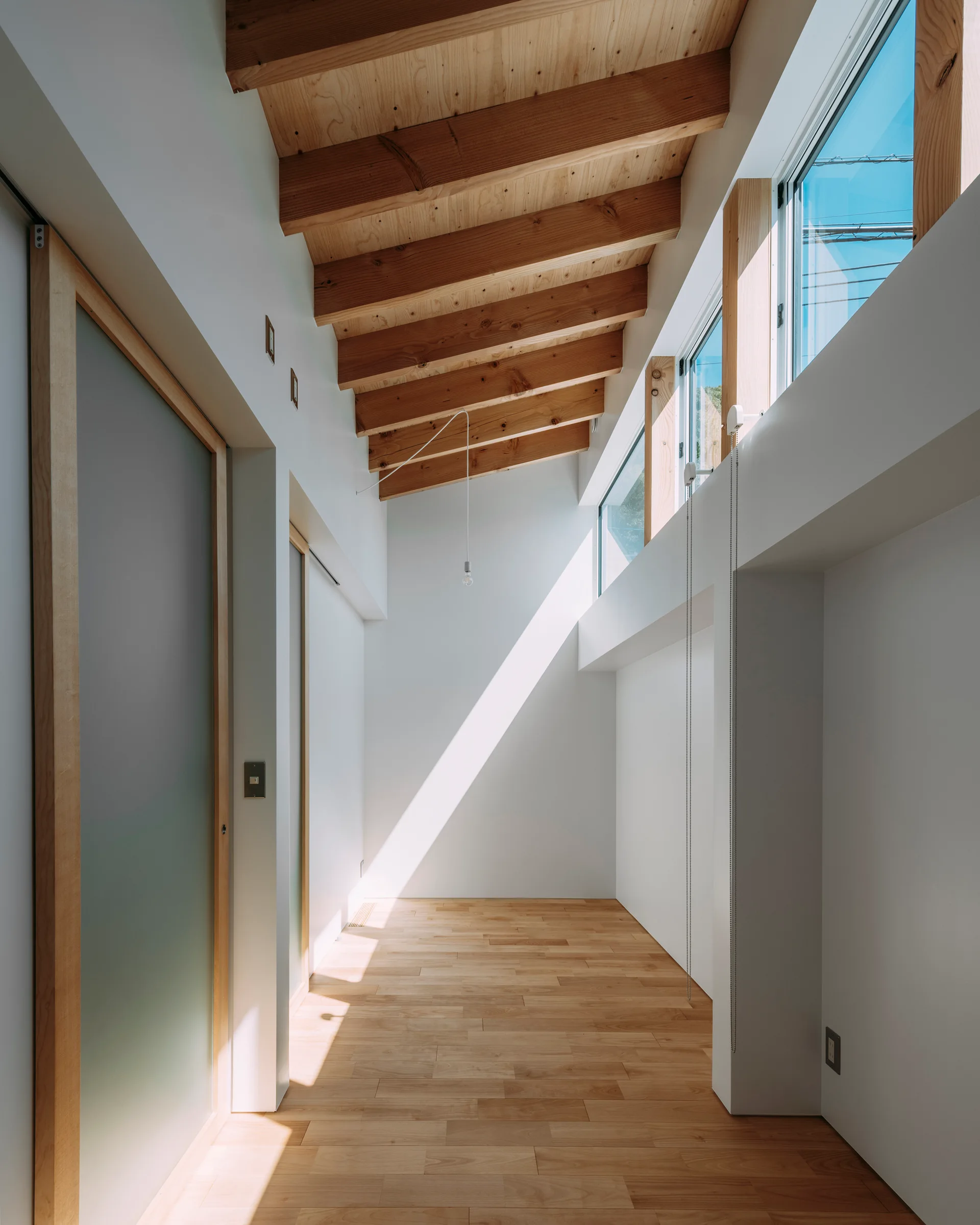 A minimalist hallway features light wood flooring, exposed wooden ceiling beams, white walls, and clerestory windows that let in bright natural light.