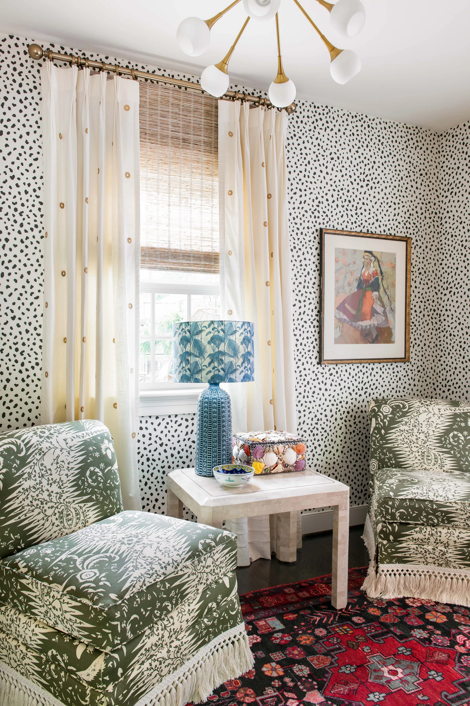 An eclectic sitting area features two green patterned armchairs with fringe trim, a white side table with a blue lamp, and black-and-white spotted wallpaper.