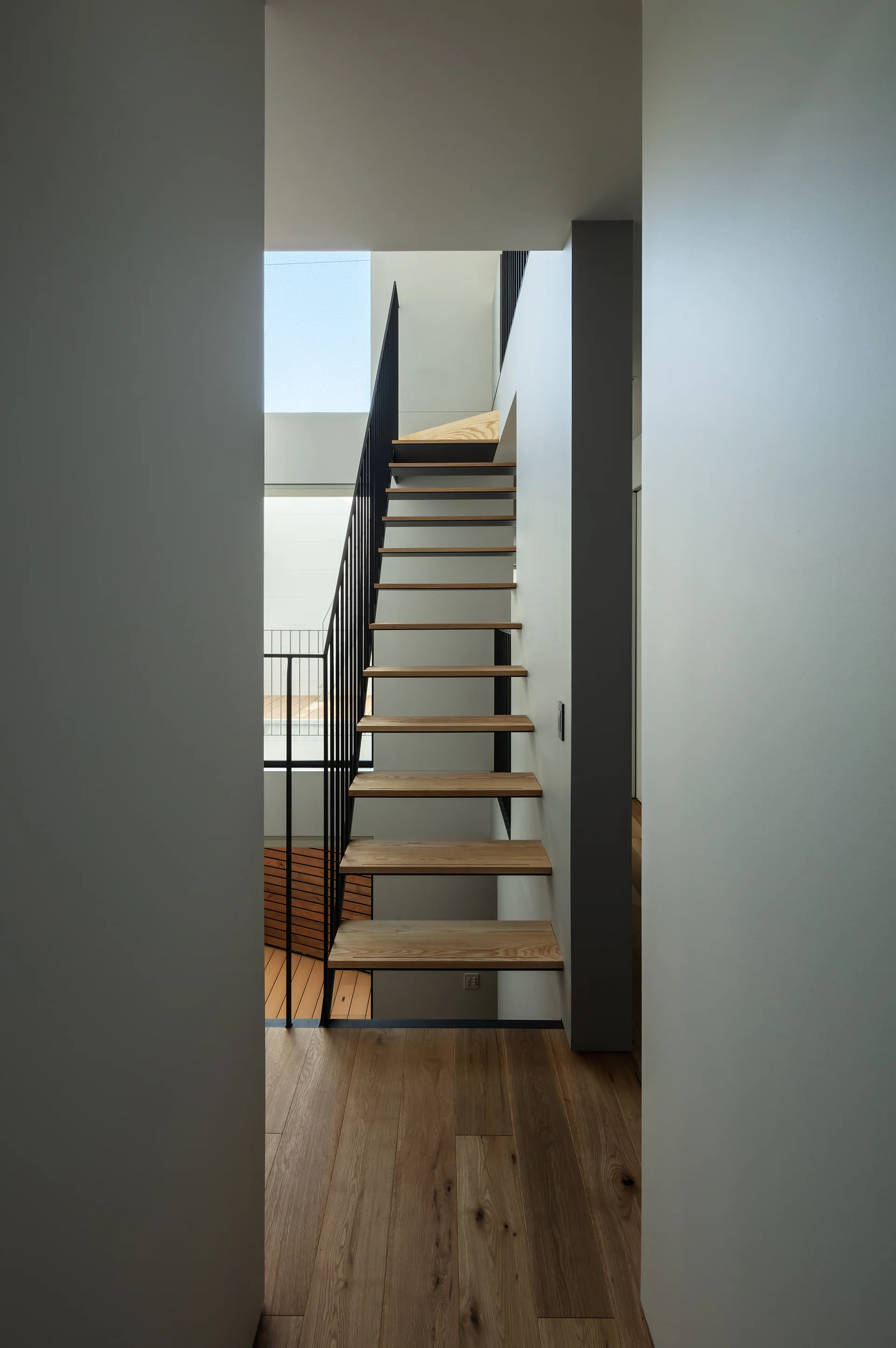 A modern floating staircase with light wood treads and vertical black metal railings is framed by white walls and illuminated by a skylight.