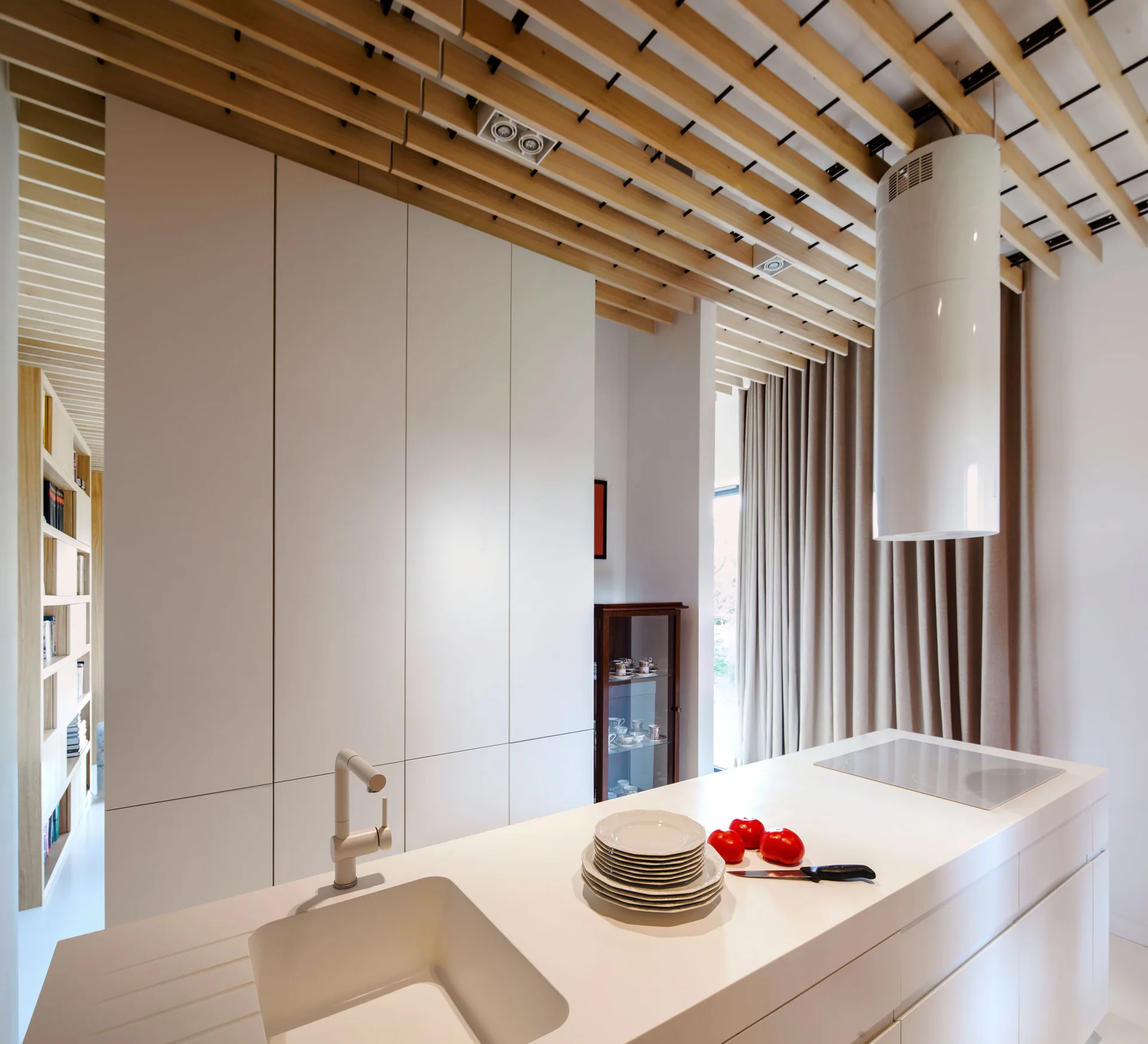 A minimalist white kitchen featuring an island with an integrated sink, seamless cabinetry, and a ceiling with light wood slats.