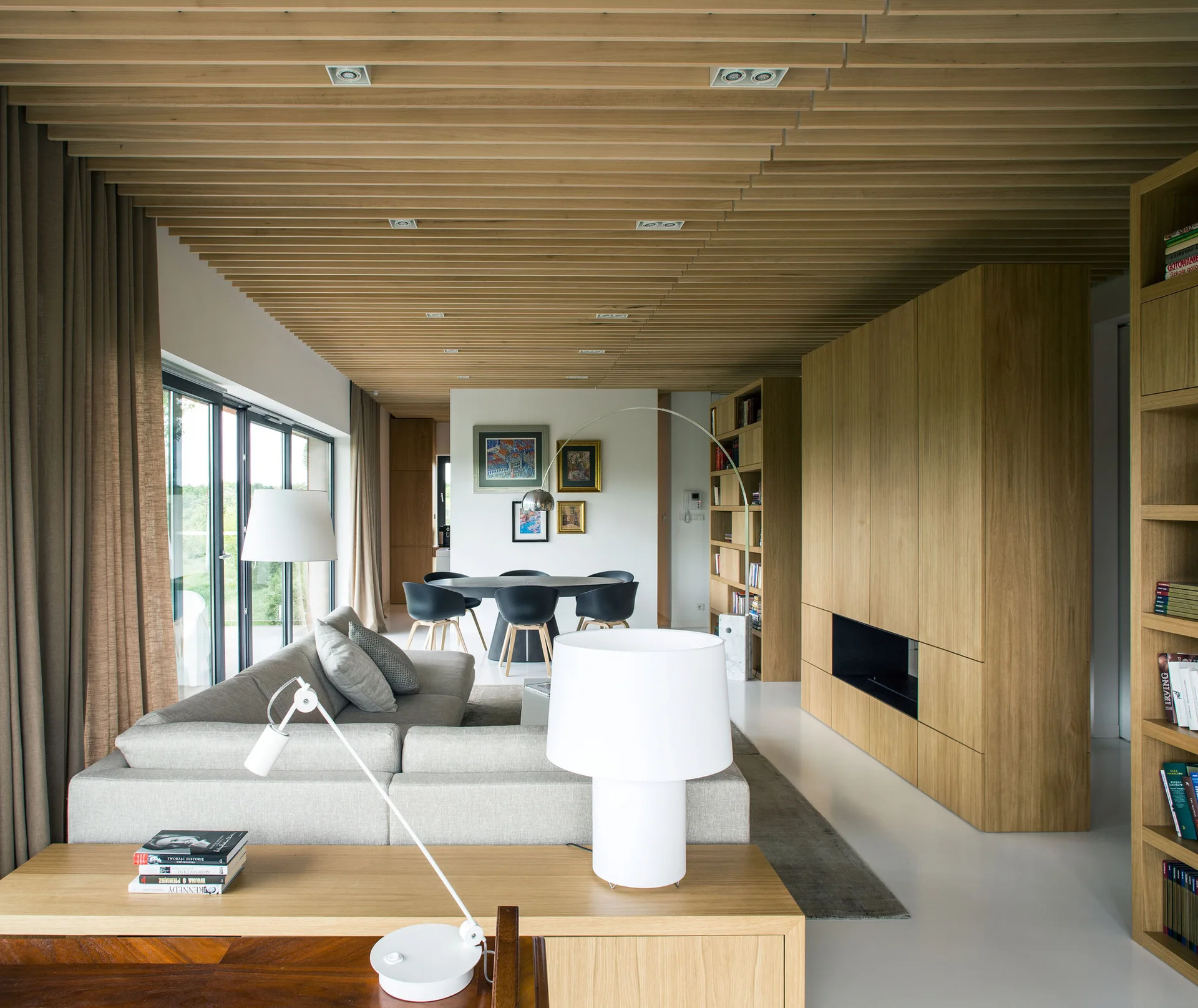 A contemporary open-plan living room with a wood slat ceiling, light gray sofa, and a large wooden media cabinet.