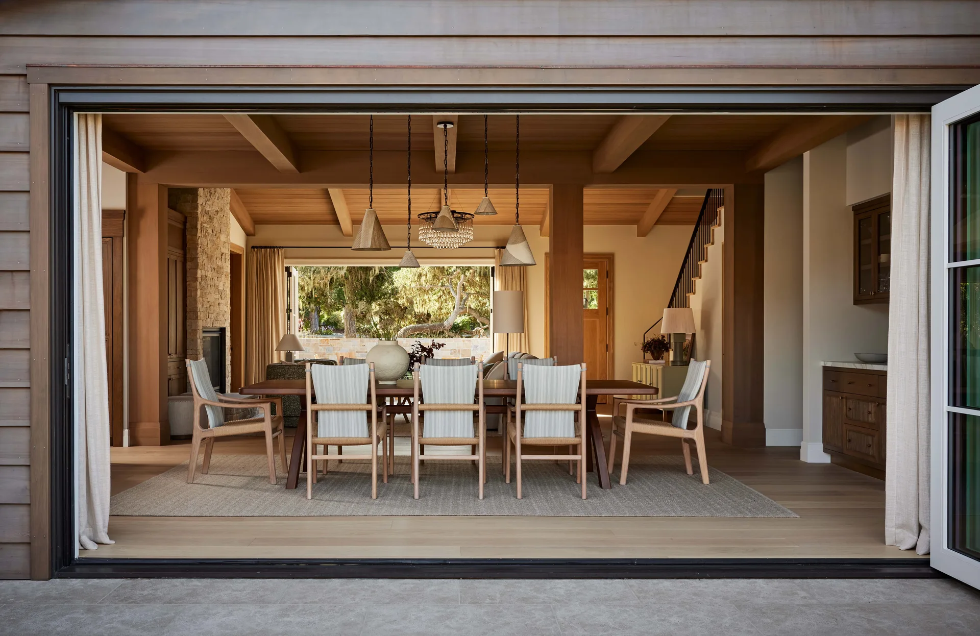 A spacious transitional dining room features a long wooden table, striped chairs, and hanging pendant lights beneath a beamed ceiling.