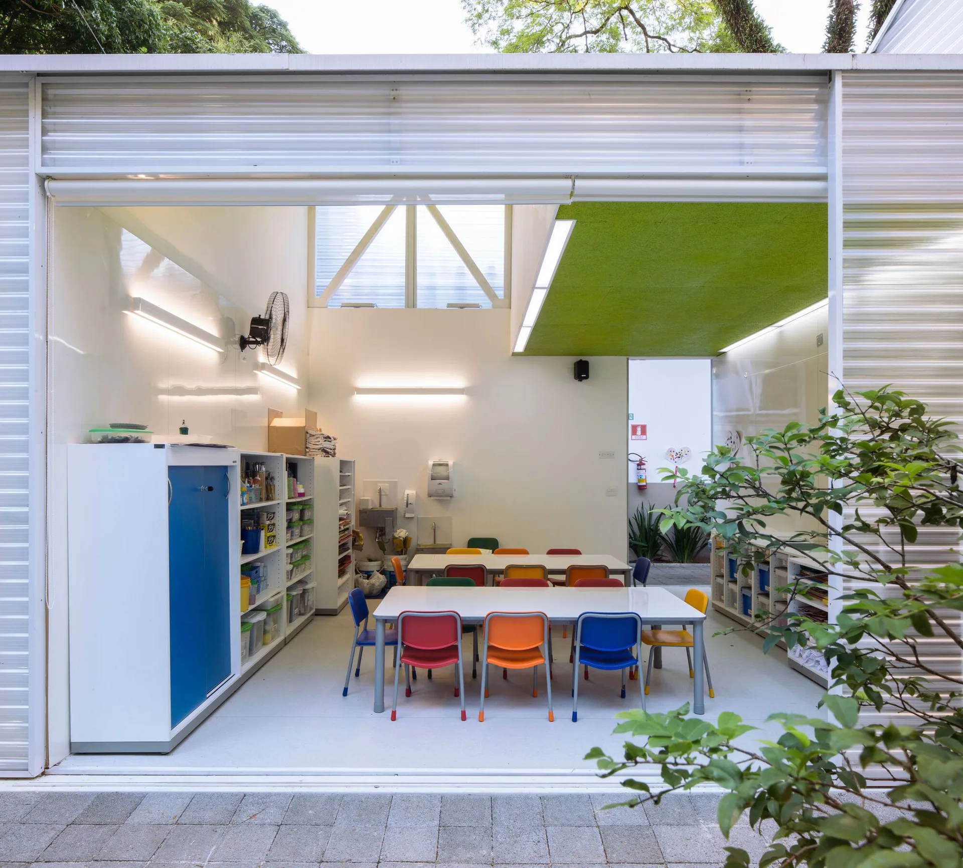An open educational studio features colorful plastic chairs at white tables, white storage shelving, and a bright green ceiling panel.