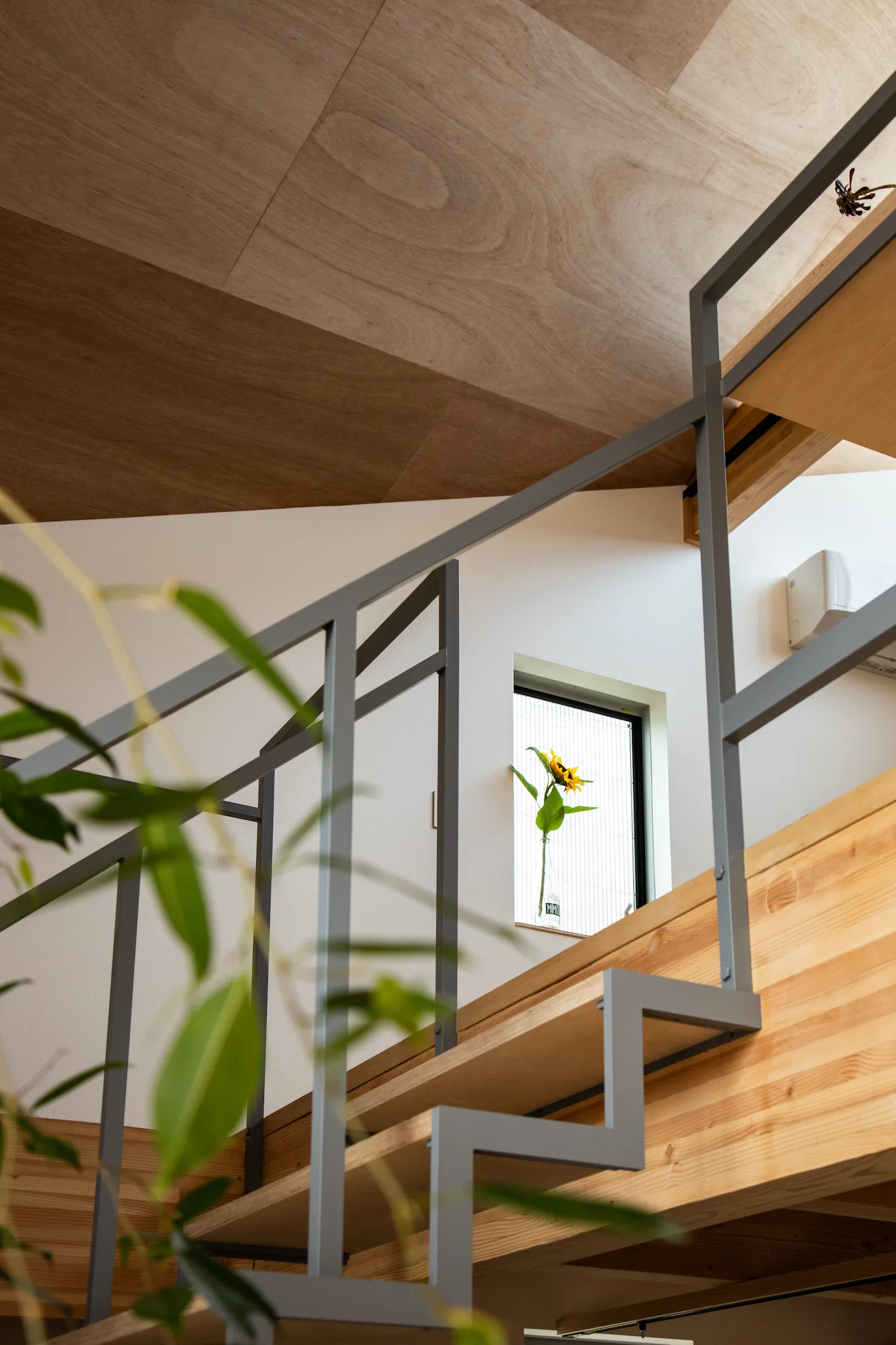 A minimalist modern staircase featuring light wood treads and a grey metal railing against white walls under a wood panel ceiling.