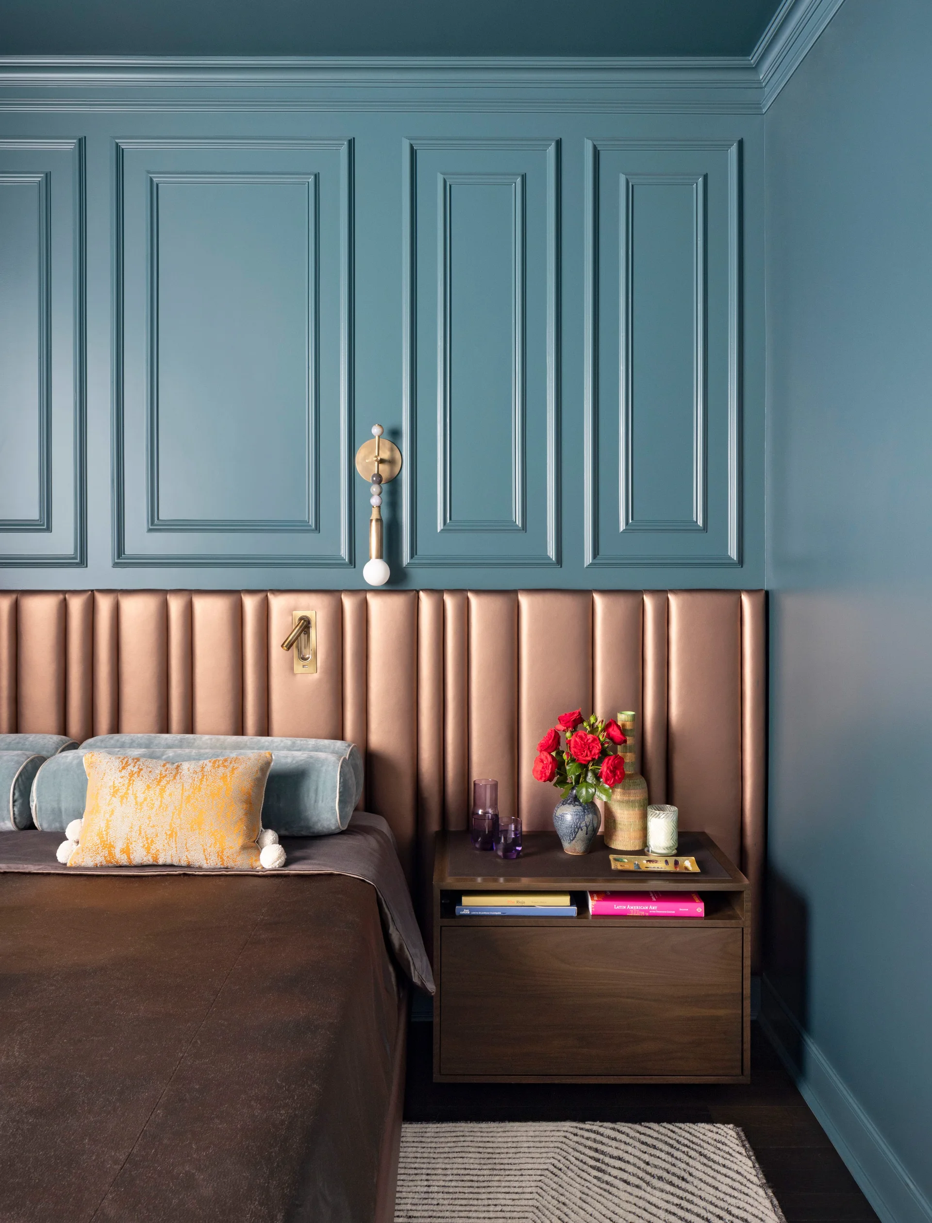 A bedroom features teal blue paneled walls, a metallic bronze channel-tufted headboard, and a dark wood nightstand with a vase of red roses.
