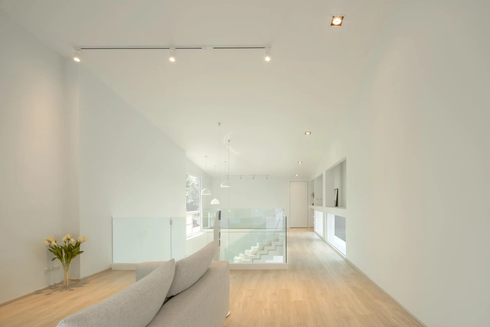 A minimalist white landing features a light grey sofa, a staircase with glass railings, and recessed niches in the wall.