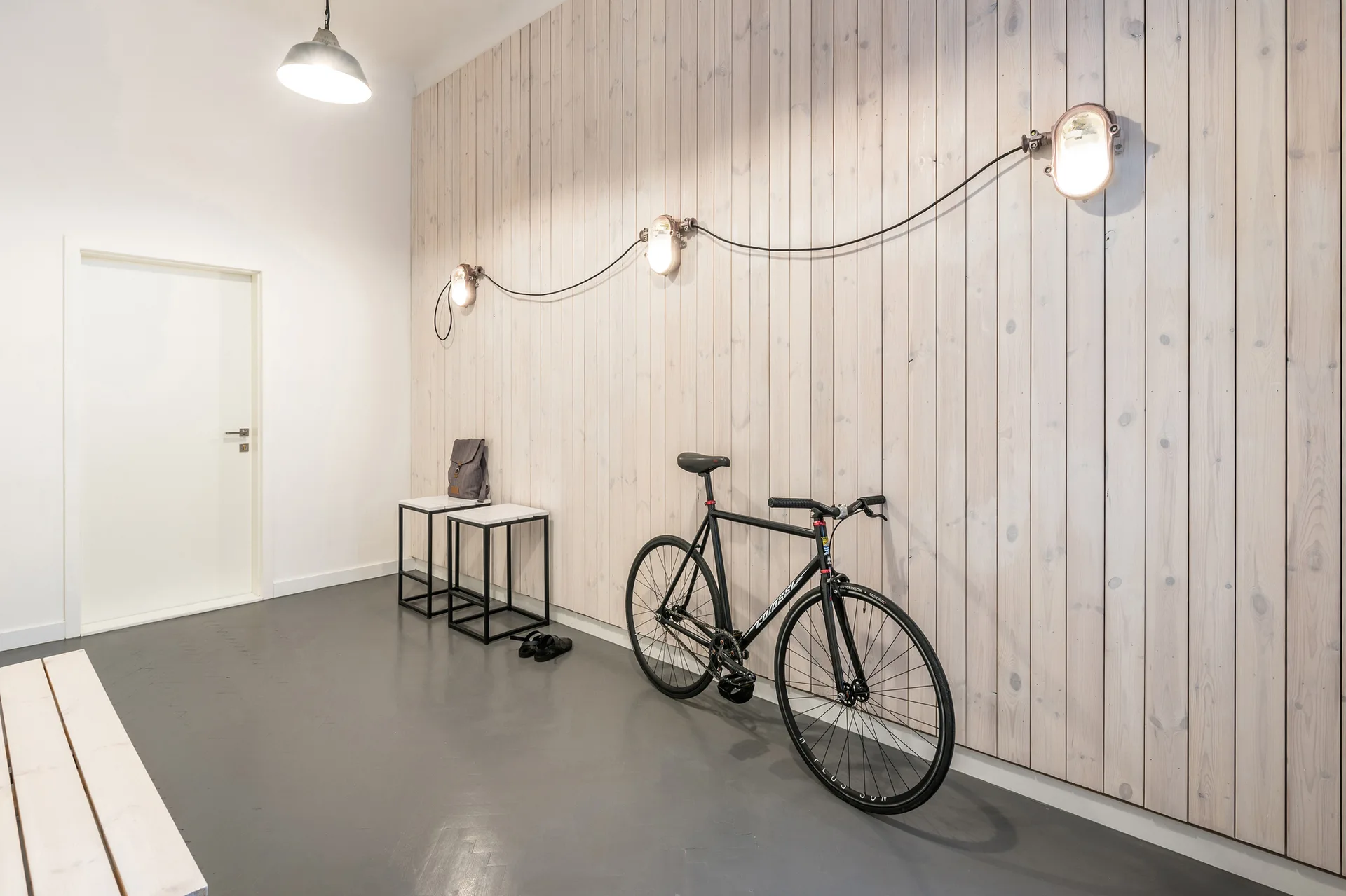 A minimalist entryway features a light wood plank accent wall, a black road bicycle, industrial wall sconces with an exposed black cord, and a grey floor.