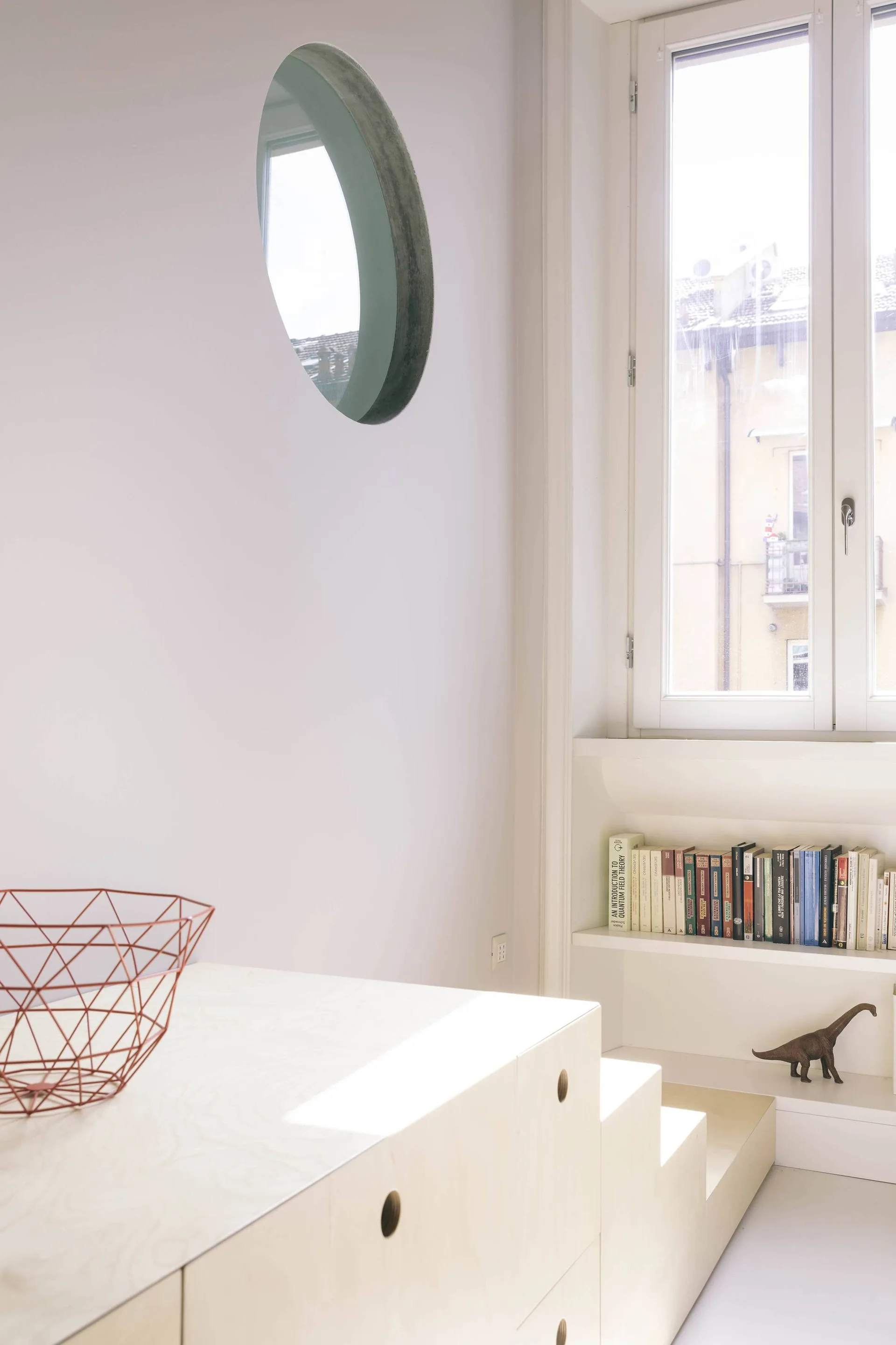 A minimalist room features a stepped plywood storage unit, a built-in bookshelf under a window, and a circular mirror on a white wall.