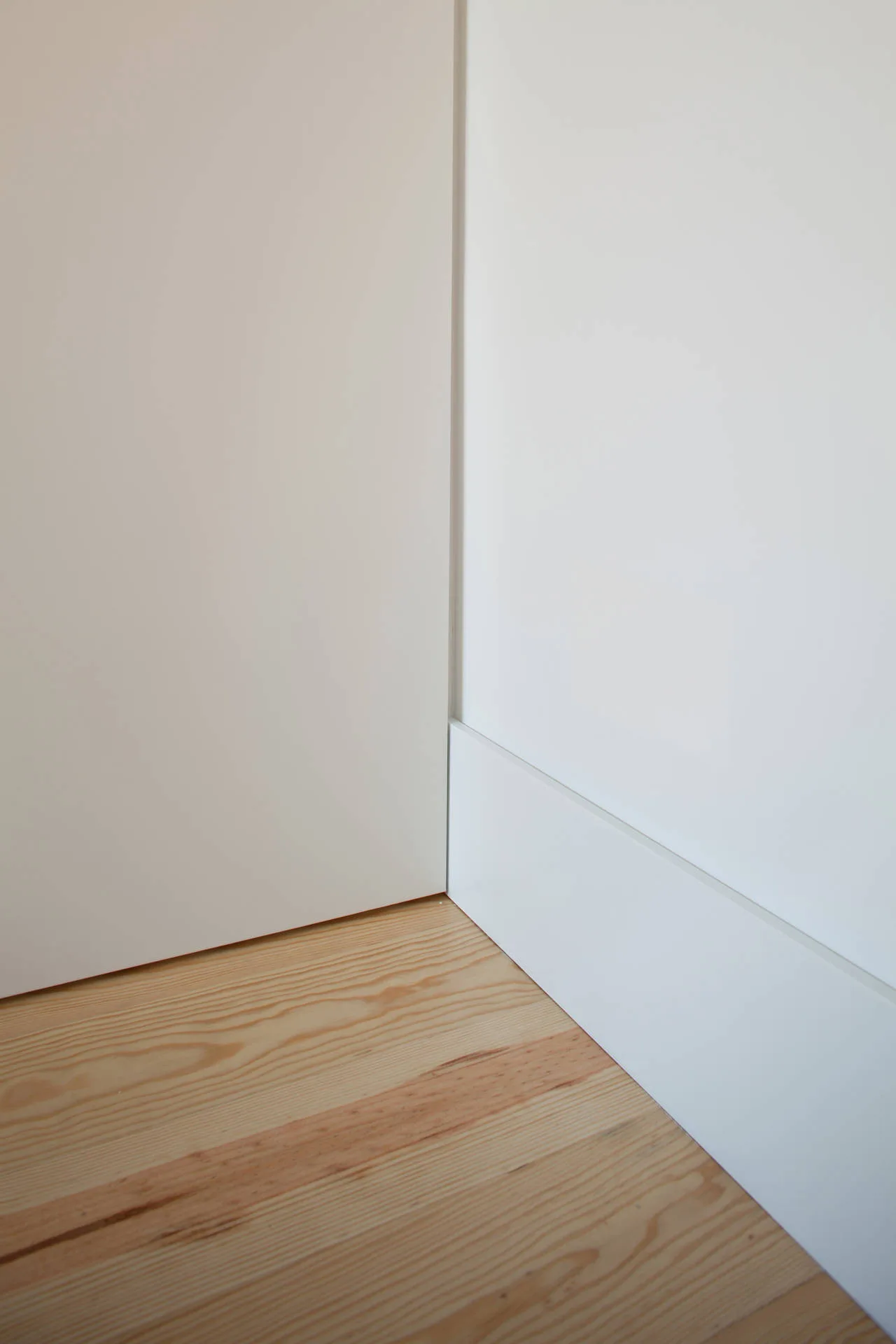 Close-up of a minimalist white painted wall corner with a flat baseboard meeting light-toned natural wood plank flooring.
