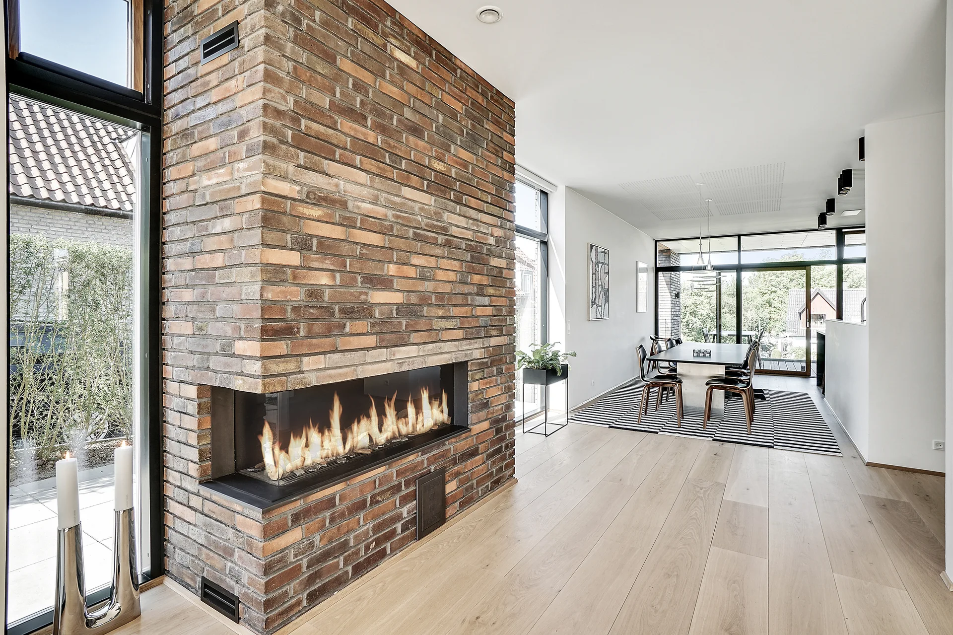 A modern open-plan dining area features a large brick chimney breast with a corner gas fireplace, light wood floors, and floor-to-ceiling windows.