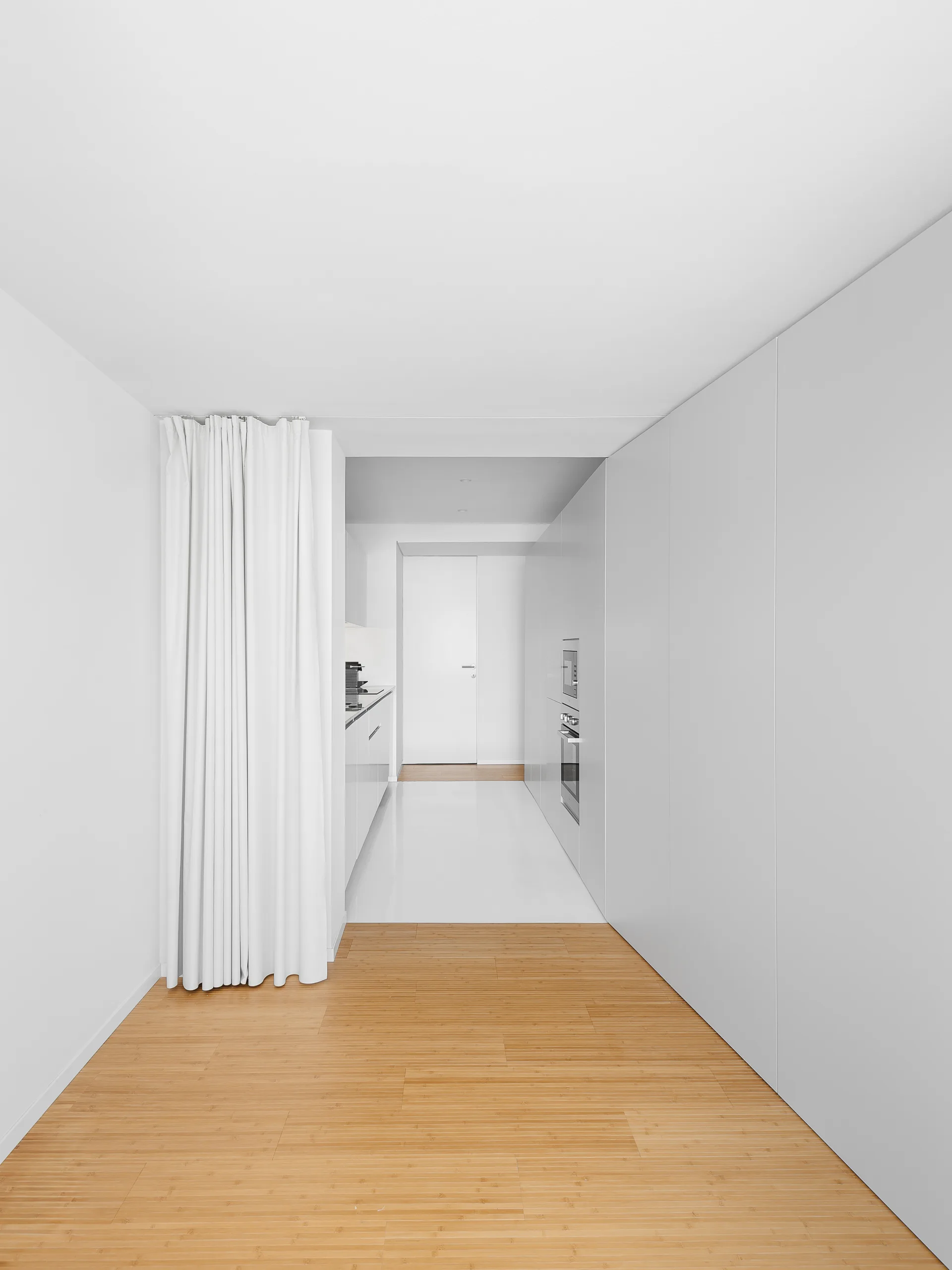 A minimalist white kitchen and hallway featuring seamless wall cabinets, integrated appliances, light wood flooring, and a white curtain partition.