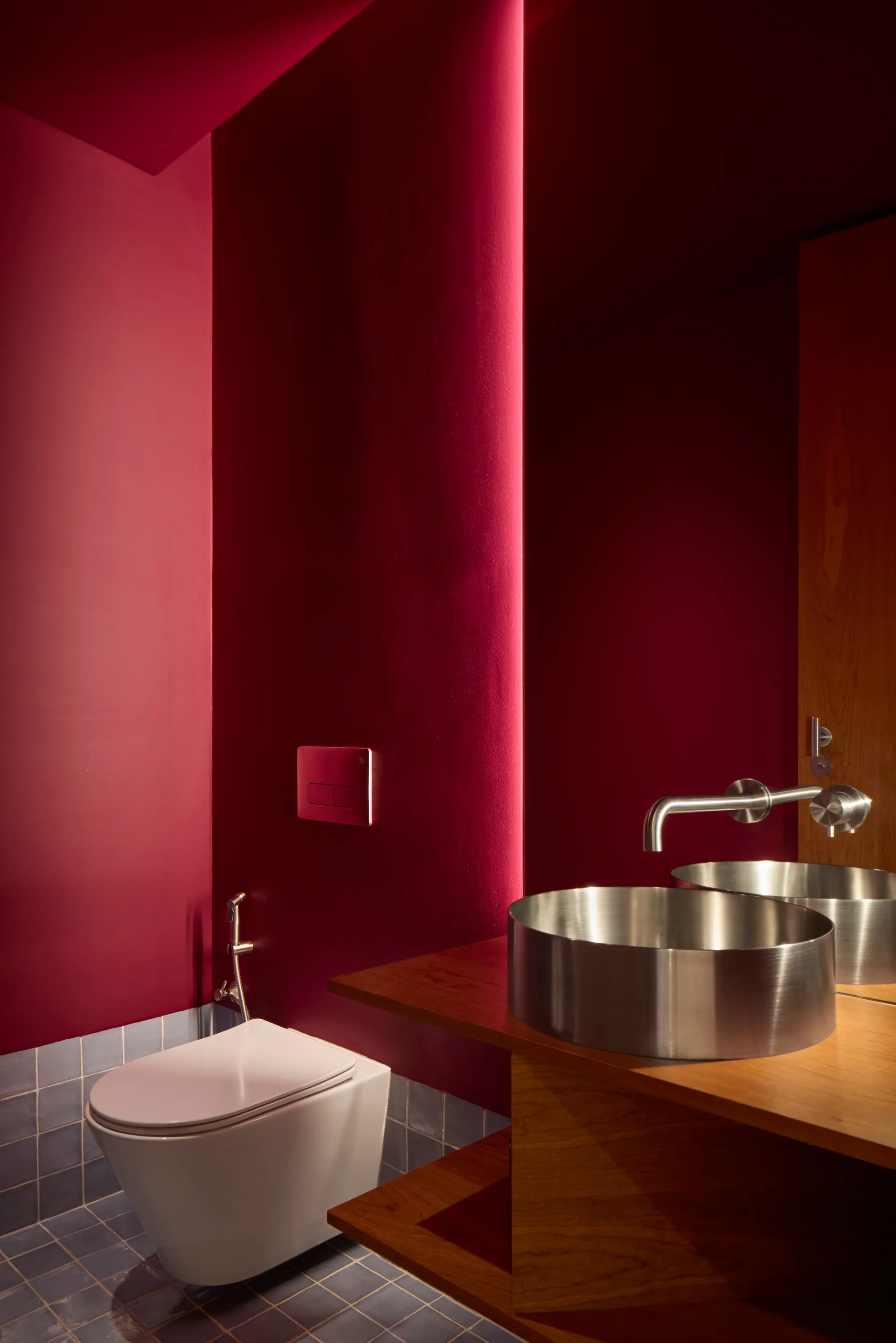 A modern powder room features deep red walls, a wall-mounted toilet, blue floor tiles, and a floating wood vanity with a round brushed metal vessel sink.