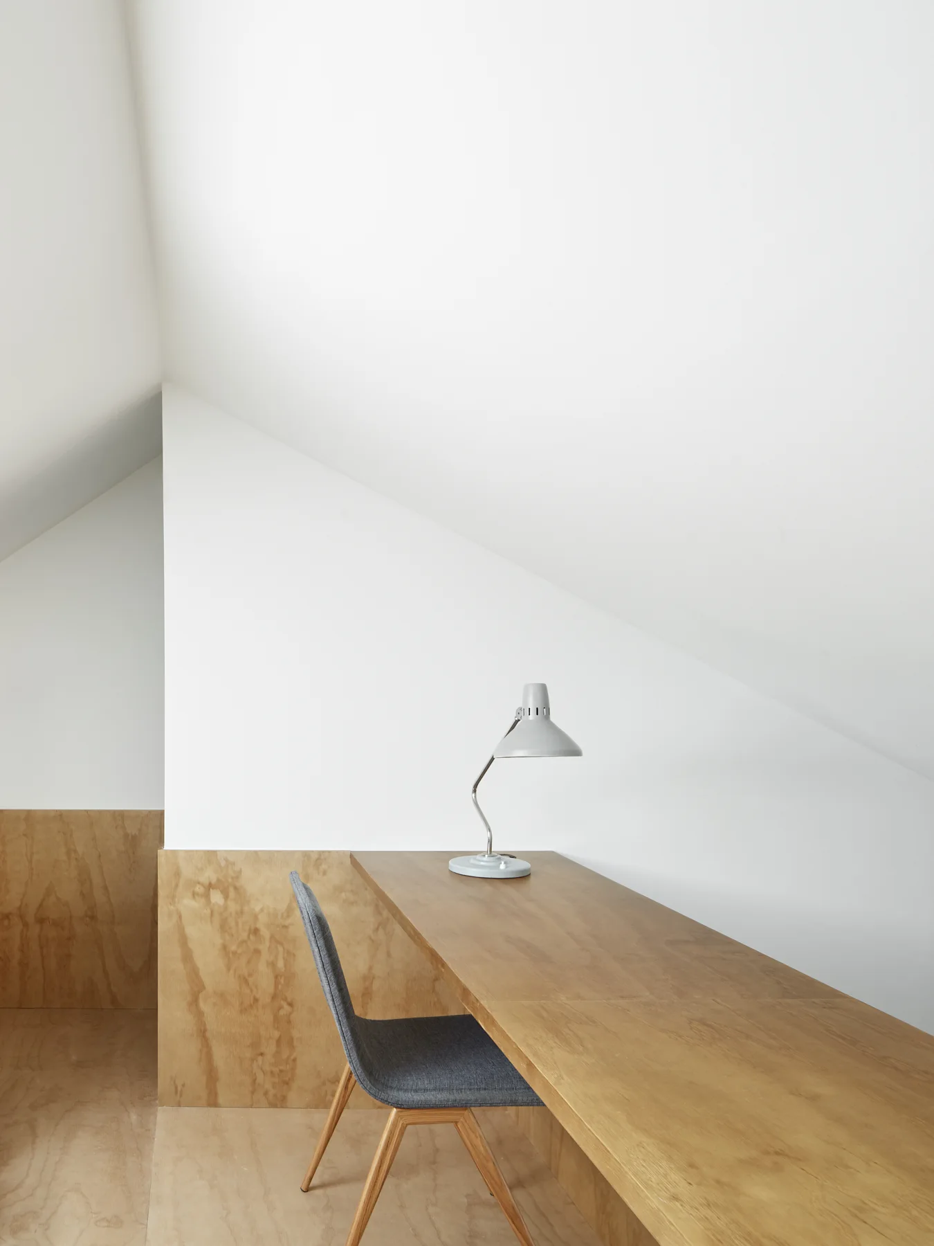 A minimalist attic workspace features a long wooden desk with a grey chair and table lamp under a slanted white ceiling.