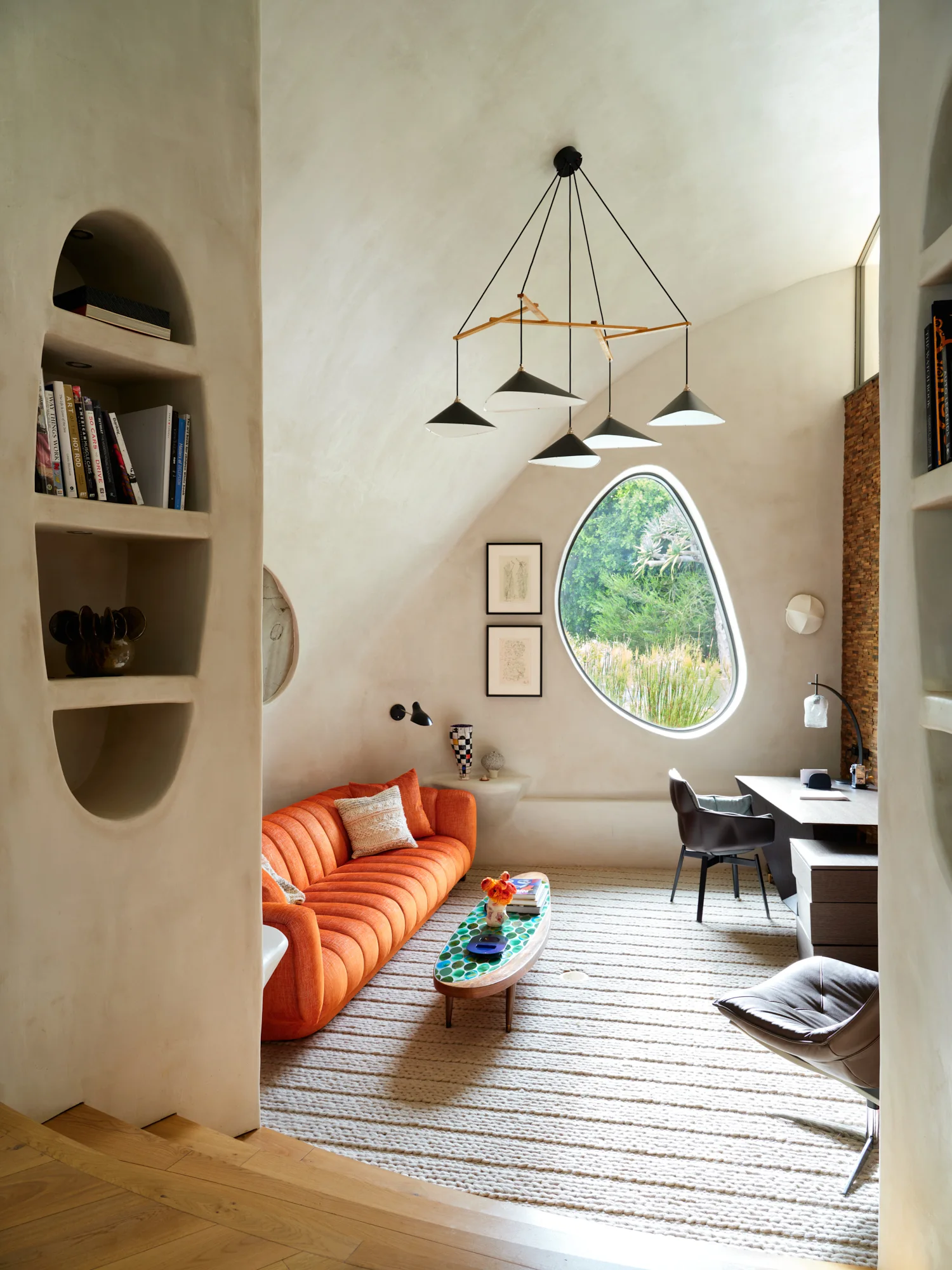 A living room with curved cream plaster walls, a bright orange channel-tufted sofa, a dark wood desk, and an organic-shaped window.