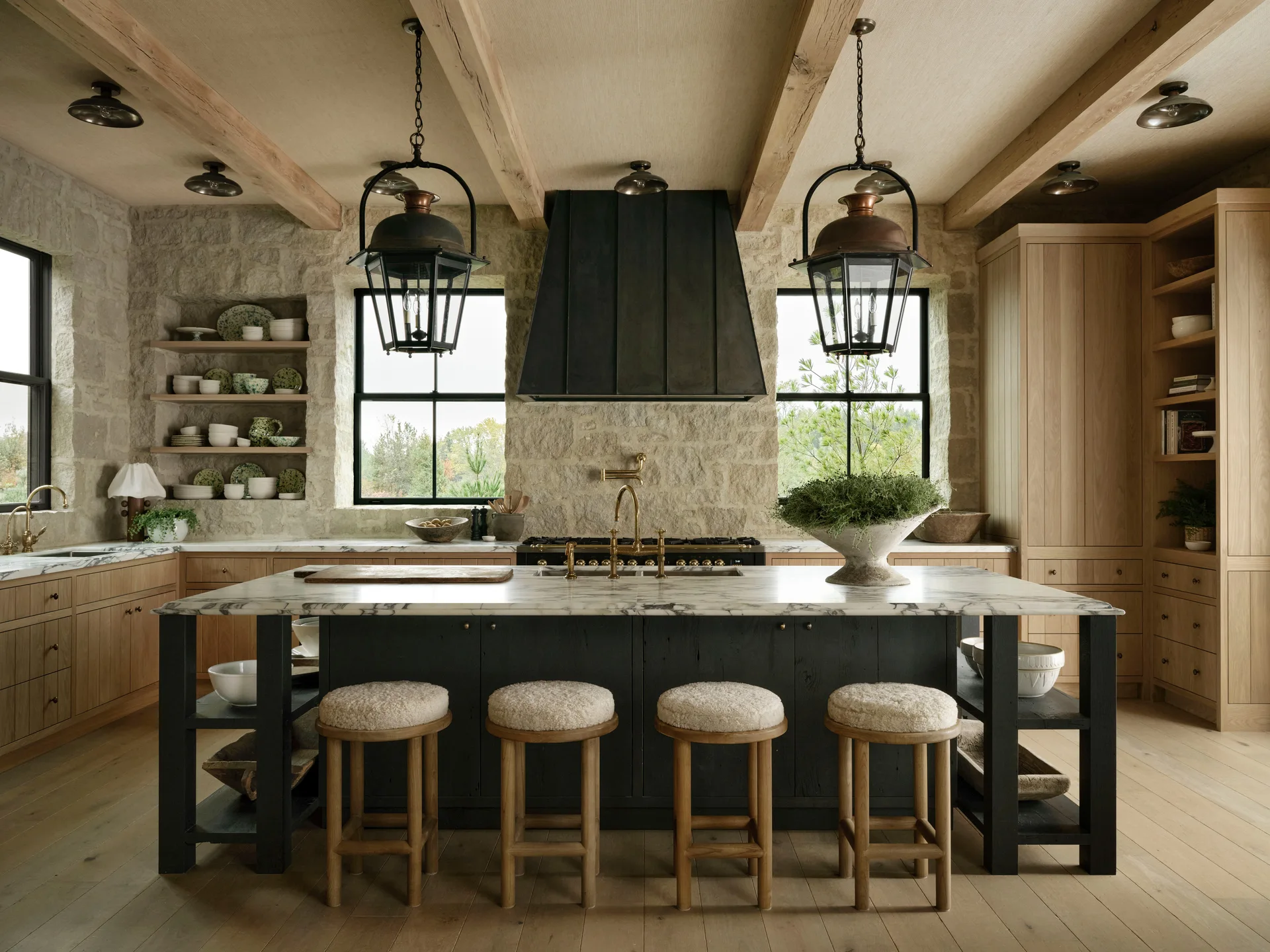 A rustic kitchen features a dark wood island with a marble countertop, four textured stools, wooden ceiling beams, and a natural stone backsplash wall.