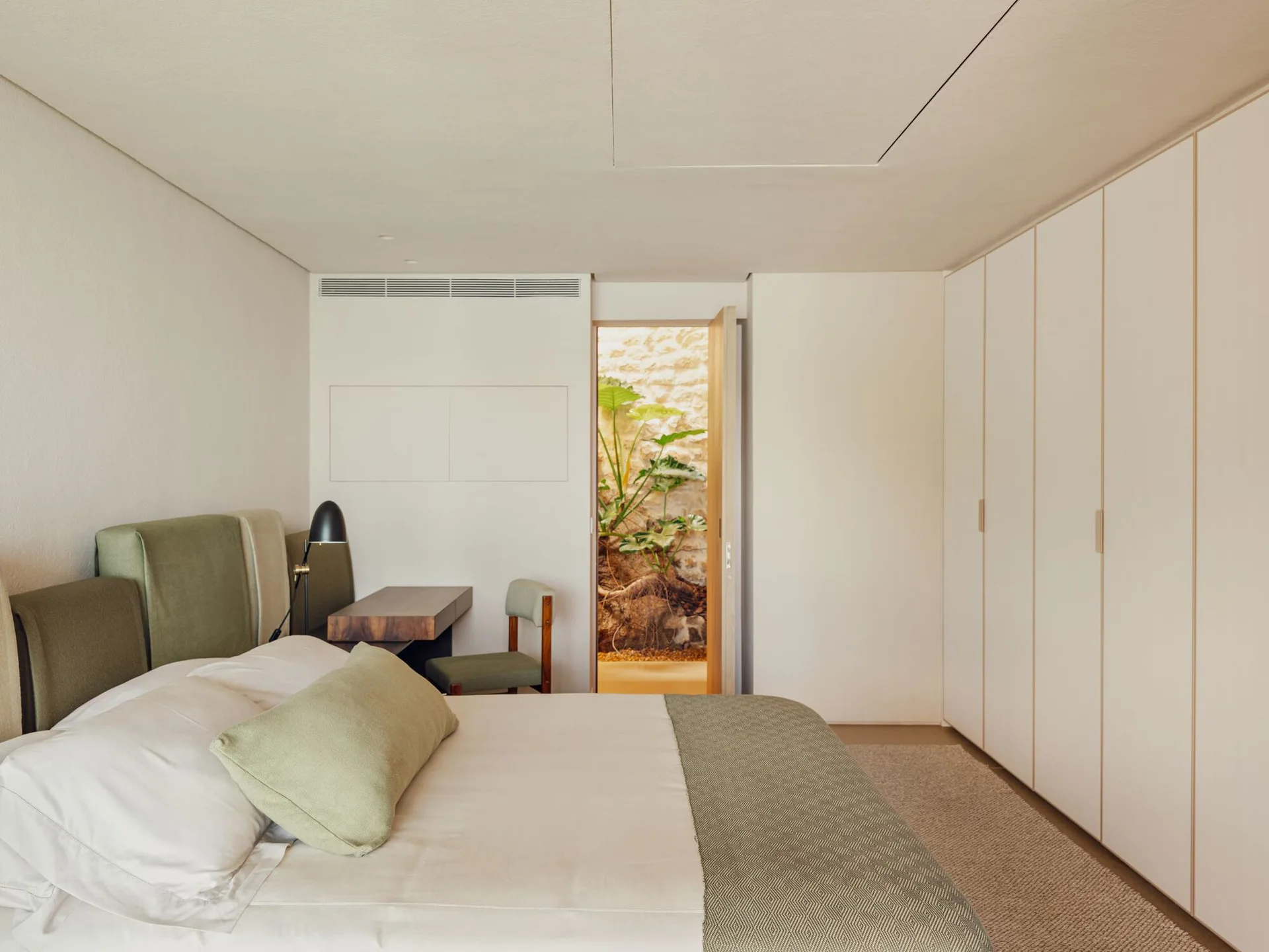 A minimalist off-white bedroom featuring an olive green upholstered bed, built-in wardrobes, and an open door looking out to a stone-walled courtyard.