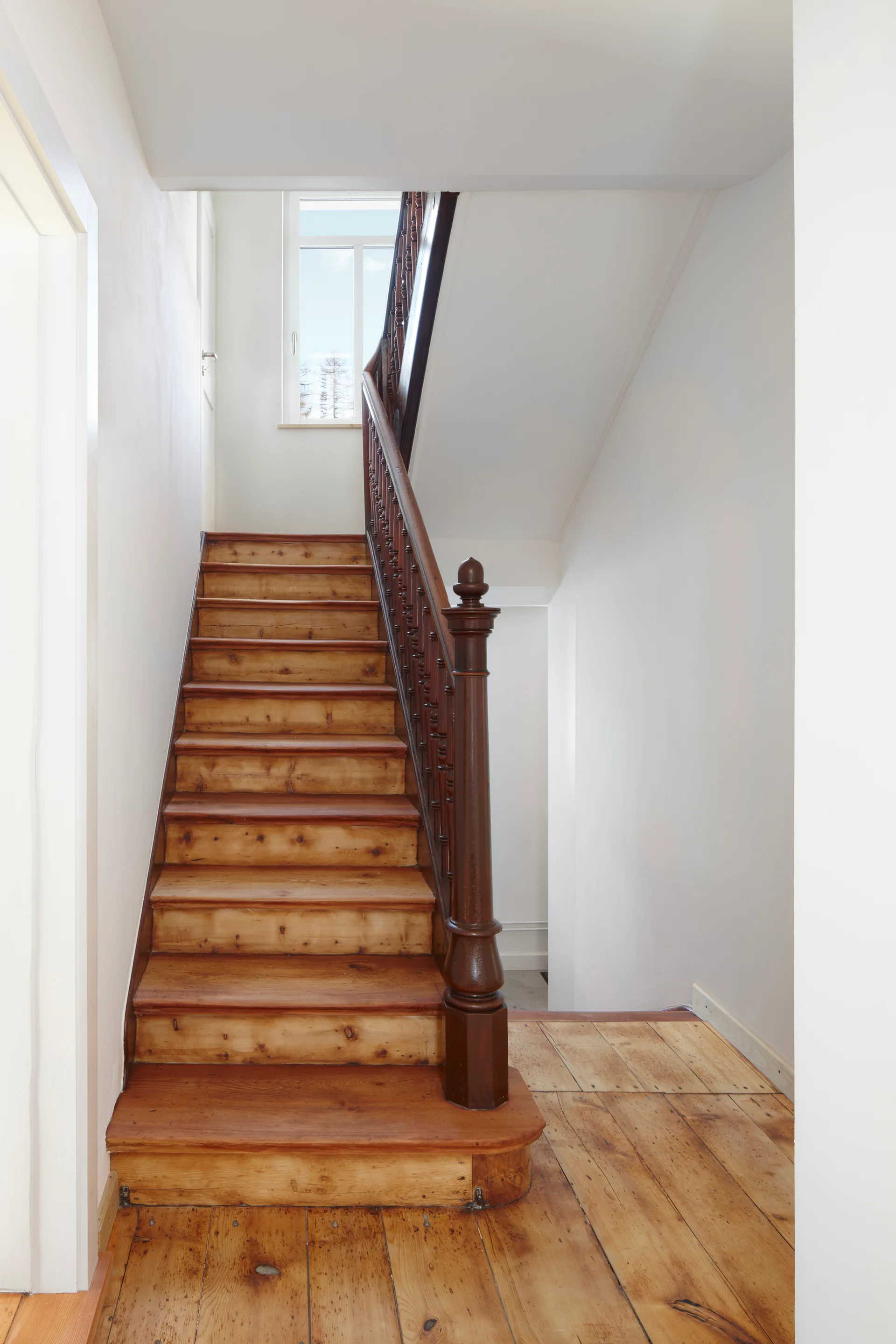 A traditional wooden staircase features light wood treads, wide plank floors, and a contrasting dark brown turned baluster railing under white walls.