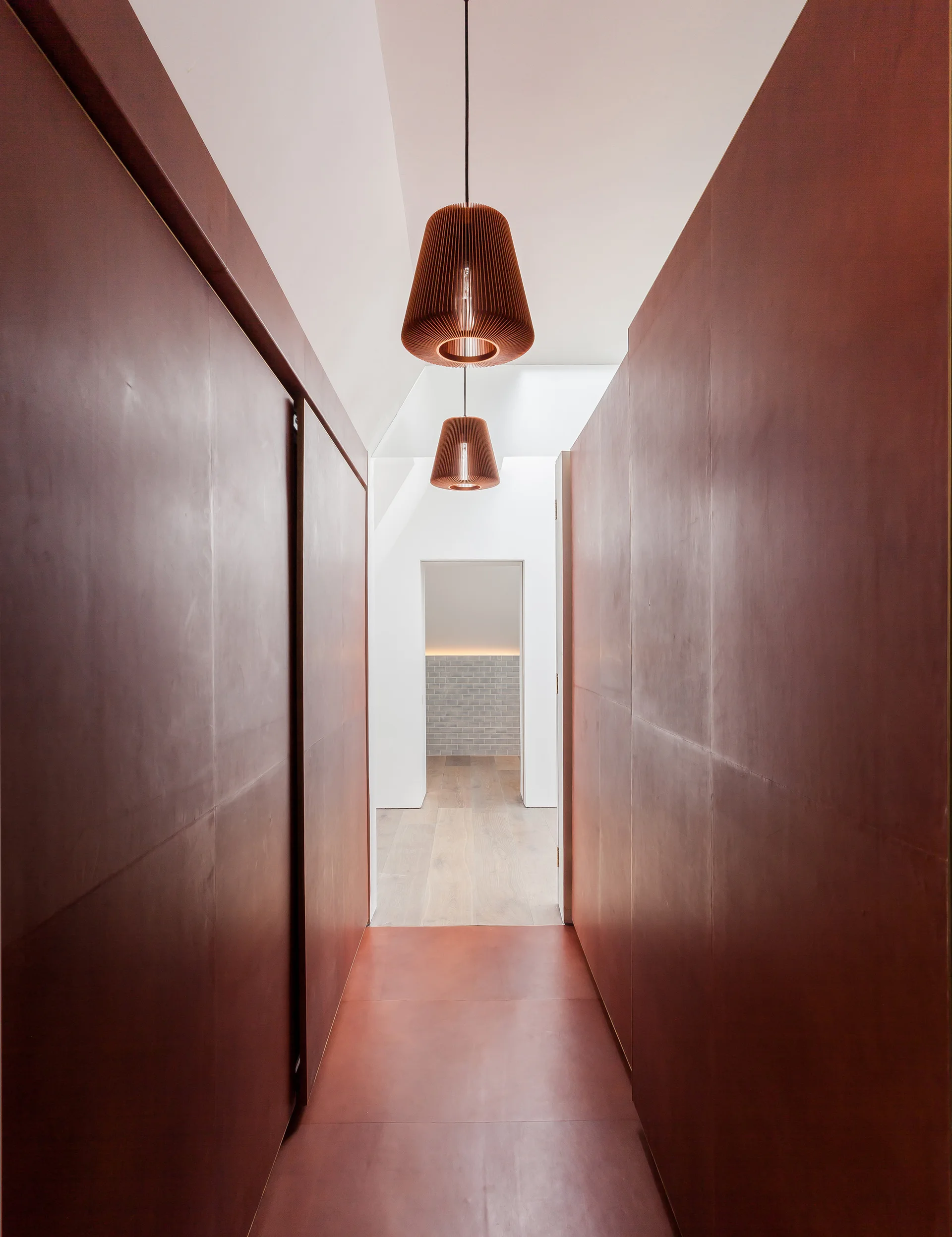 A symmetrical hallway with matte burgundy leather wall panels, matching floor tiles, and two ribbed pendant lights hanging from a white ceiling.